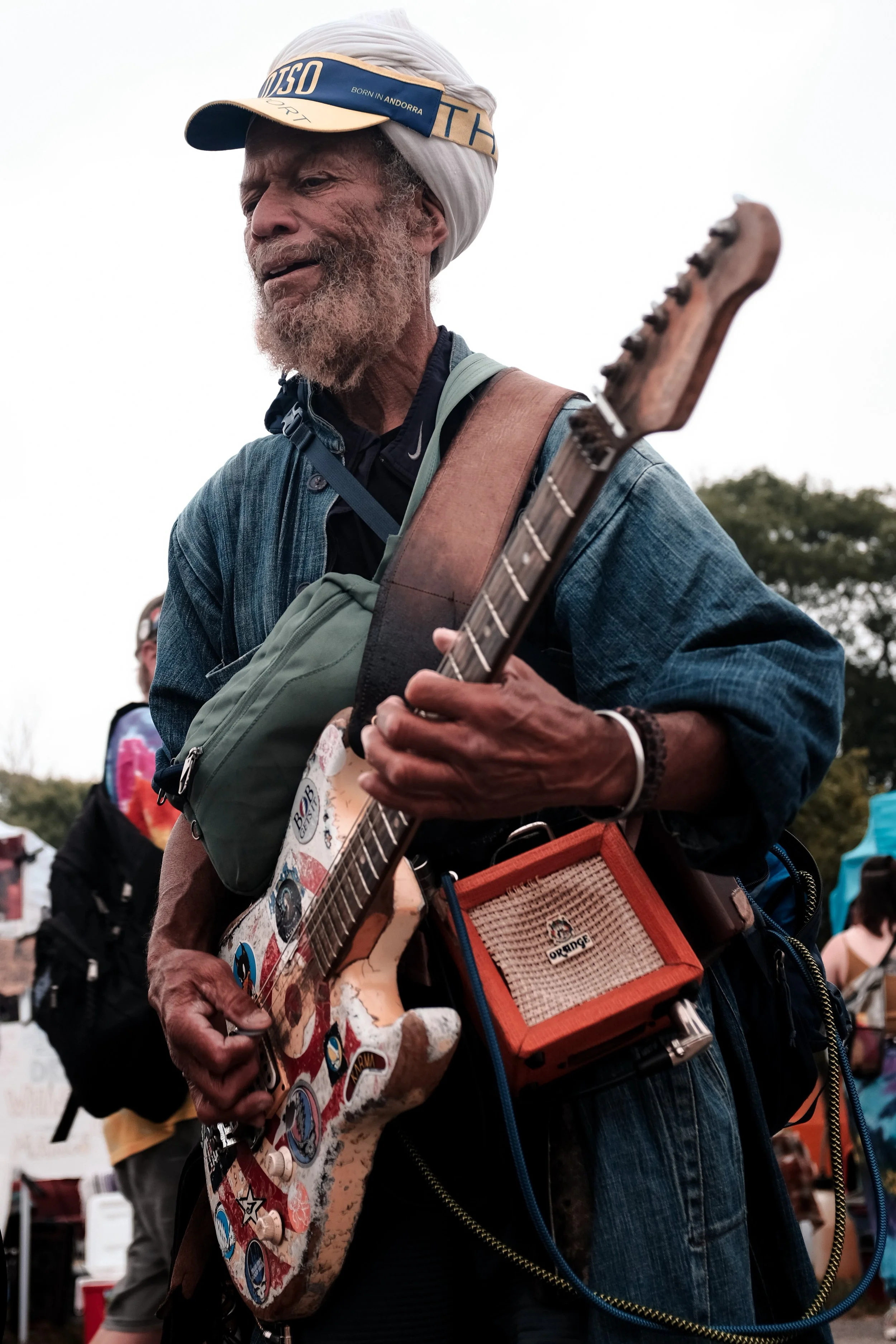 An elderly man with a white turban, wearing a denim jacket and a visor, playing an electric guitar decorated with stickers at an outdoor event.