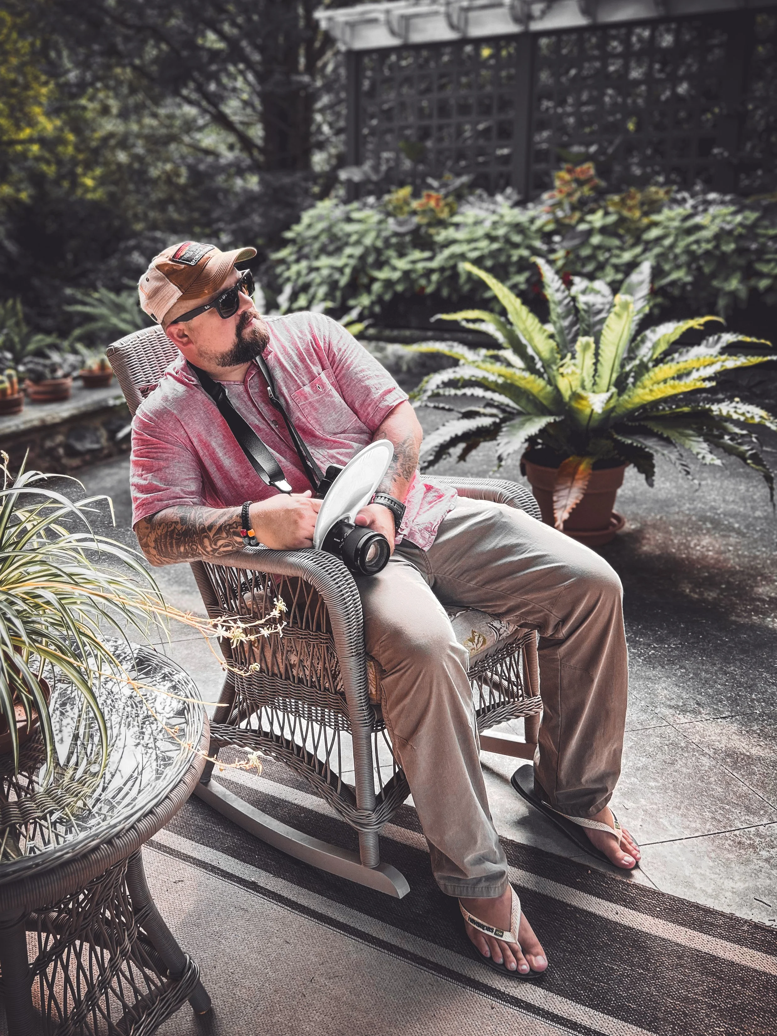A man with a beard wearing sunglasses, a beige cap, a pink shirt, khaki pants, and flip-flops is sitting in a wicker chair outdoors, holding a camera with a strap around his neck, next to a large potted plant with long green and white striped leaves.