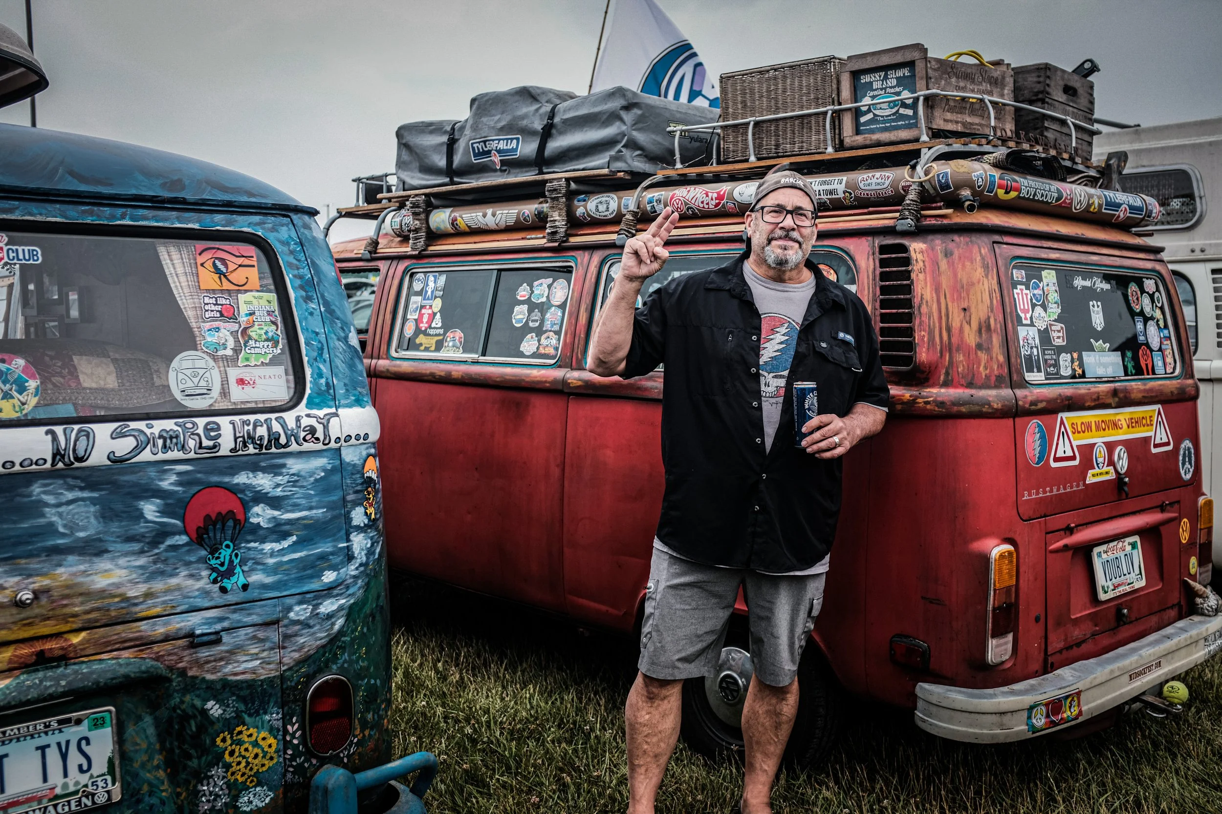 A man with glasses and a bearded face standing outdoors next to a vintage red camper van covered with stickers. The camper van has a roof rack carrying some bags and boxes. Another partially visible vehicle covered in colorful paint and stickers is n