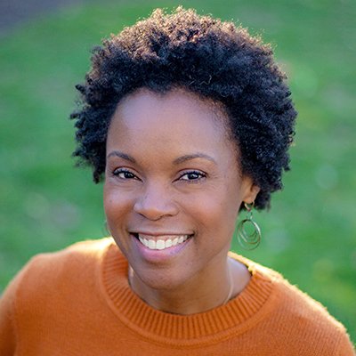 A woman with short, curly hair smiling outdoors, wearing an orange sweater against a green background.