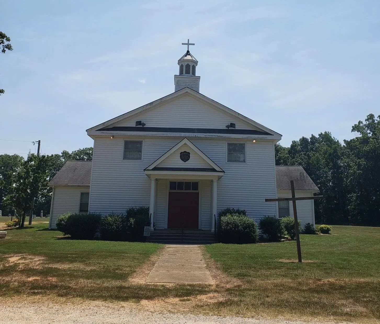 Front view of a white church with a cross on top, surrounded by green bushes and trees, with a dirt pathway leading to the entrance.