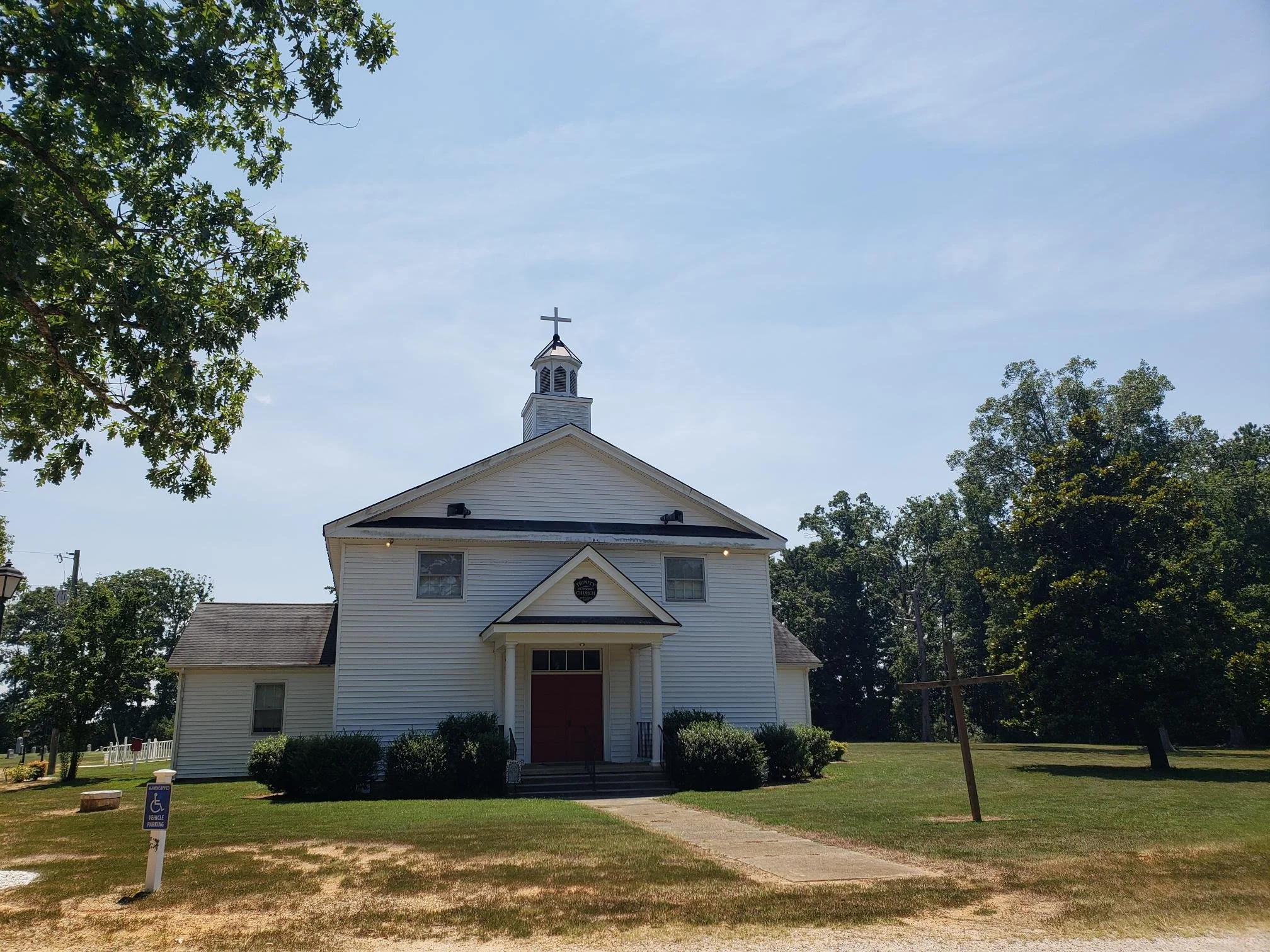 White church with a small steeple topped with a cross, set against a blue sky with trees on either side, front yard with bushes and a sidewalk, a handicap parking sign in the foreground.