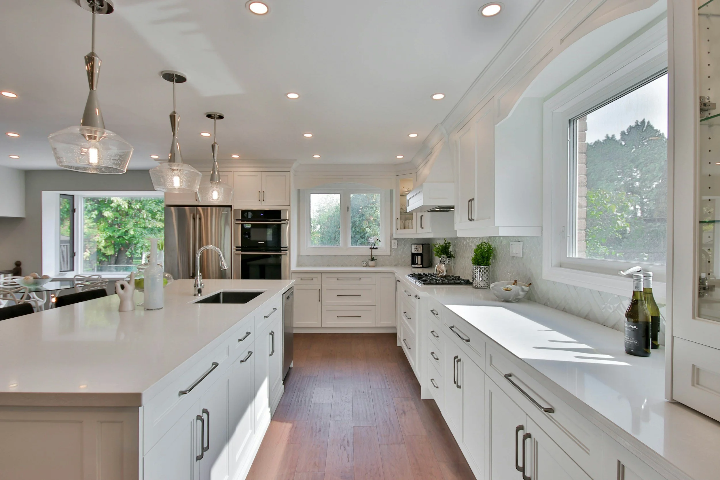 A bright, modern kitchen with white cabinets and a large white island, featuring pendant lights above. There are windows letting in natural light, and the kitchen is decorated with potted plants and bottles on the countertops.