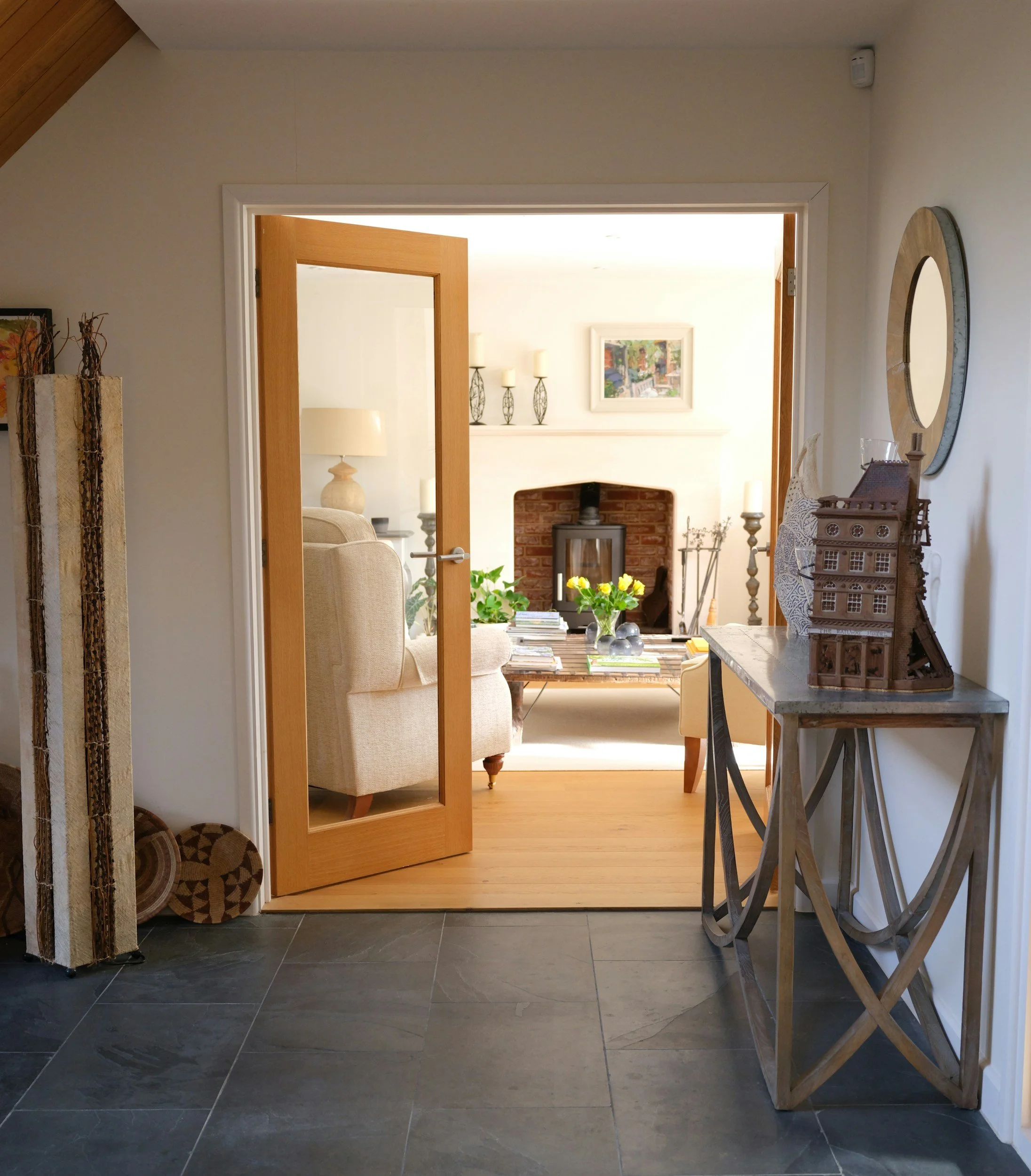 View of a living room through a wooden door with a glass panel, featuring a fireplace, artwork, candle holders, a table with books and flowers, and various decorative items.