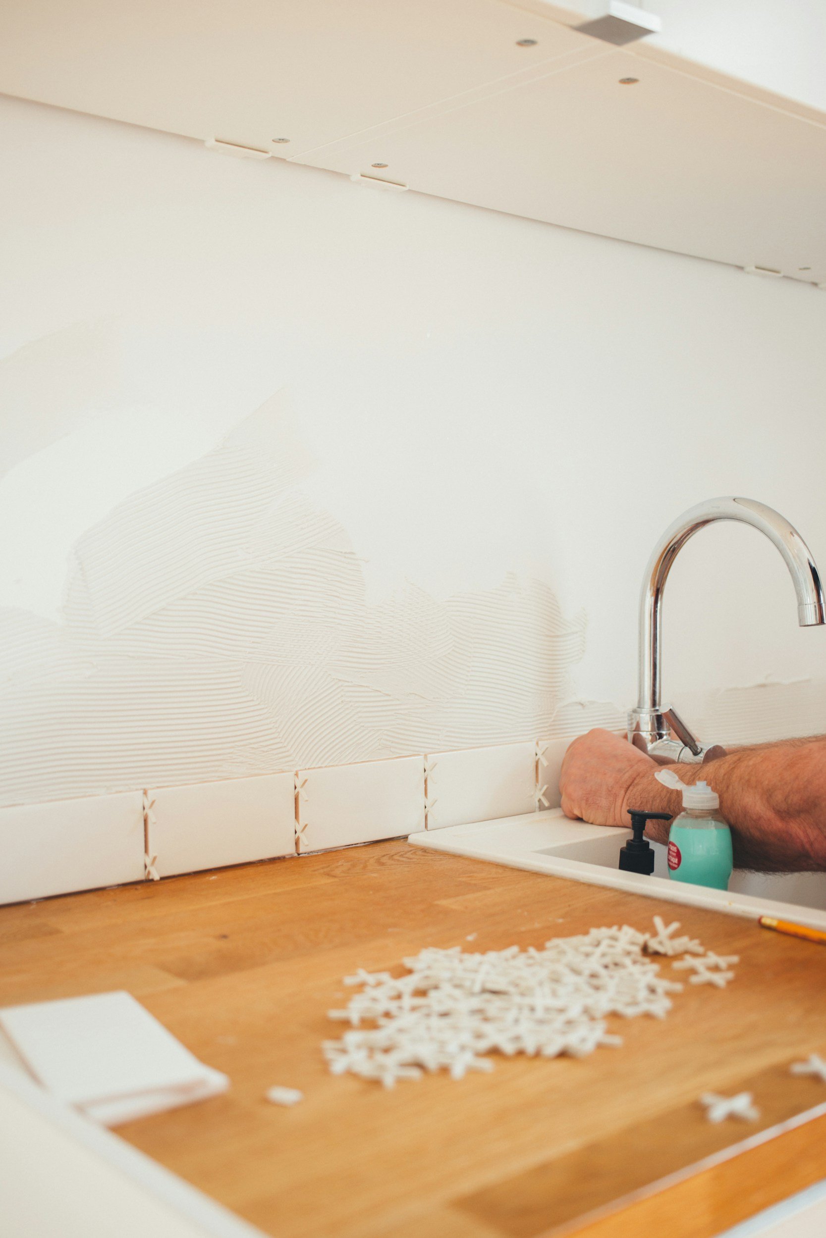 Person working on creating a tile mosaic backsplash above a kitchen sink, with white subway tiles and a small pile of mosaic tiles on the counter.