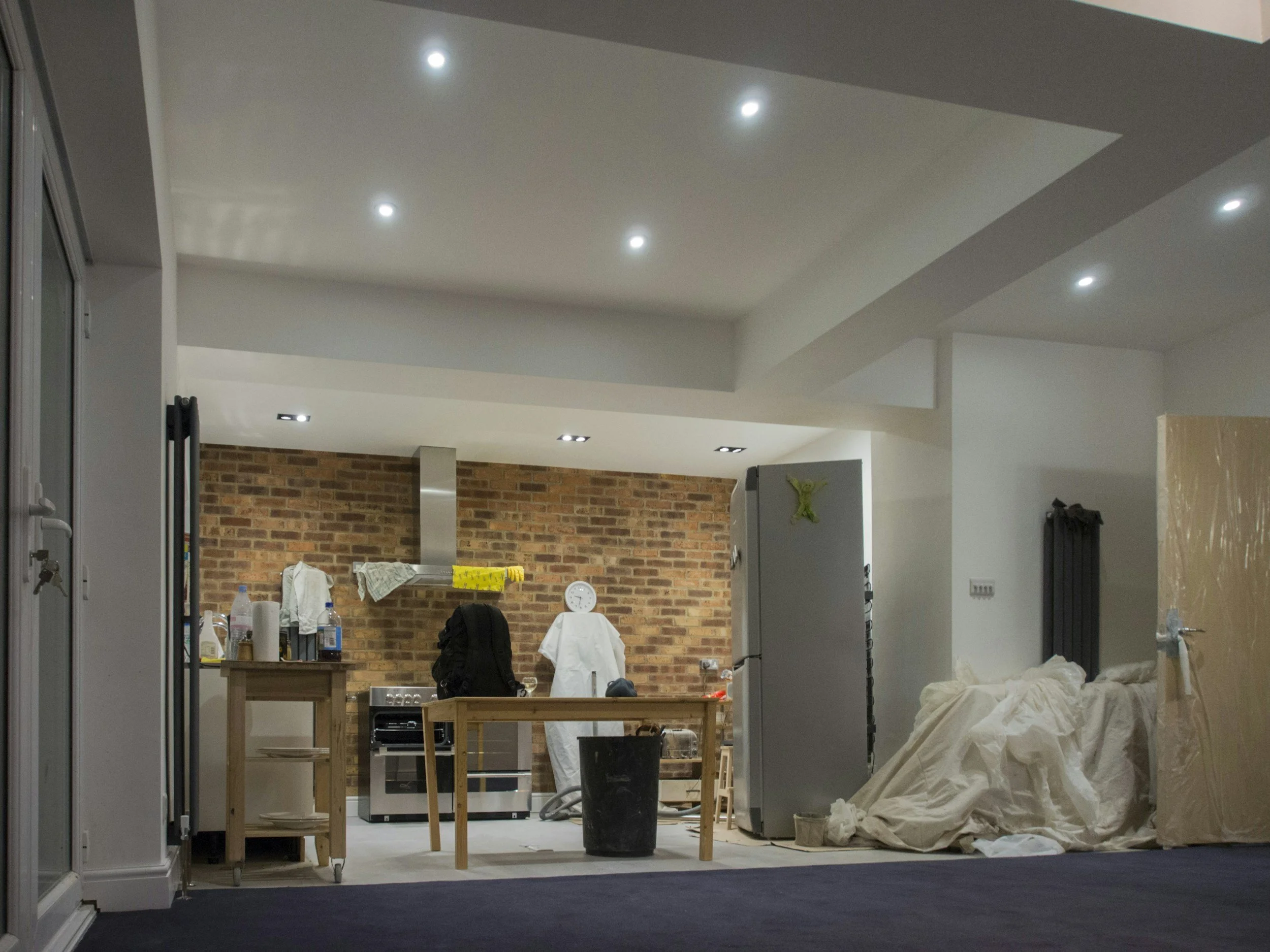 An indoor space under construction or renovation, showing wooden tables, a black trash bin, a packed refrigerator, brick wall, white cloth or tarp, and various household or renovation tools and items scattered around.