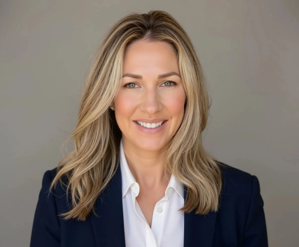 Headshot of a smiling blonde woman with wavy hair, wearing a dark blazer and white shirt, against a neutral background.