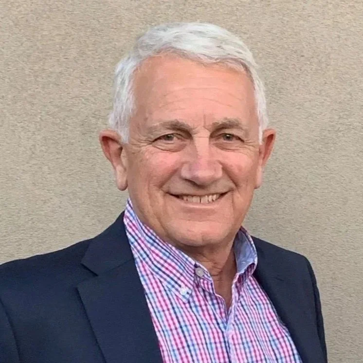 An older man with white hair smiling, wearing a navy blazer and a red and blue checkered shirt, standing against a beige wall.