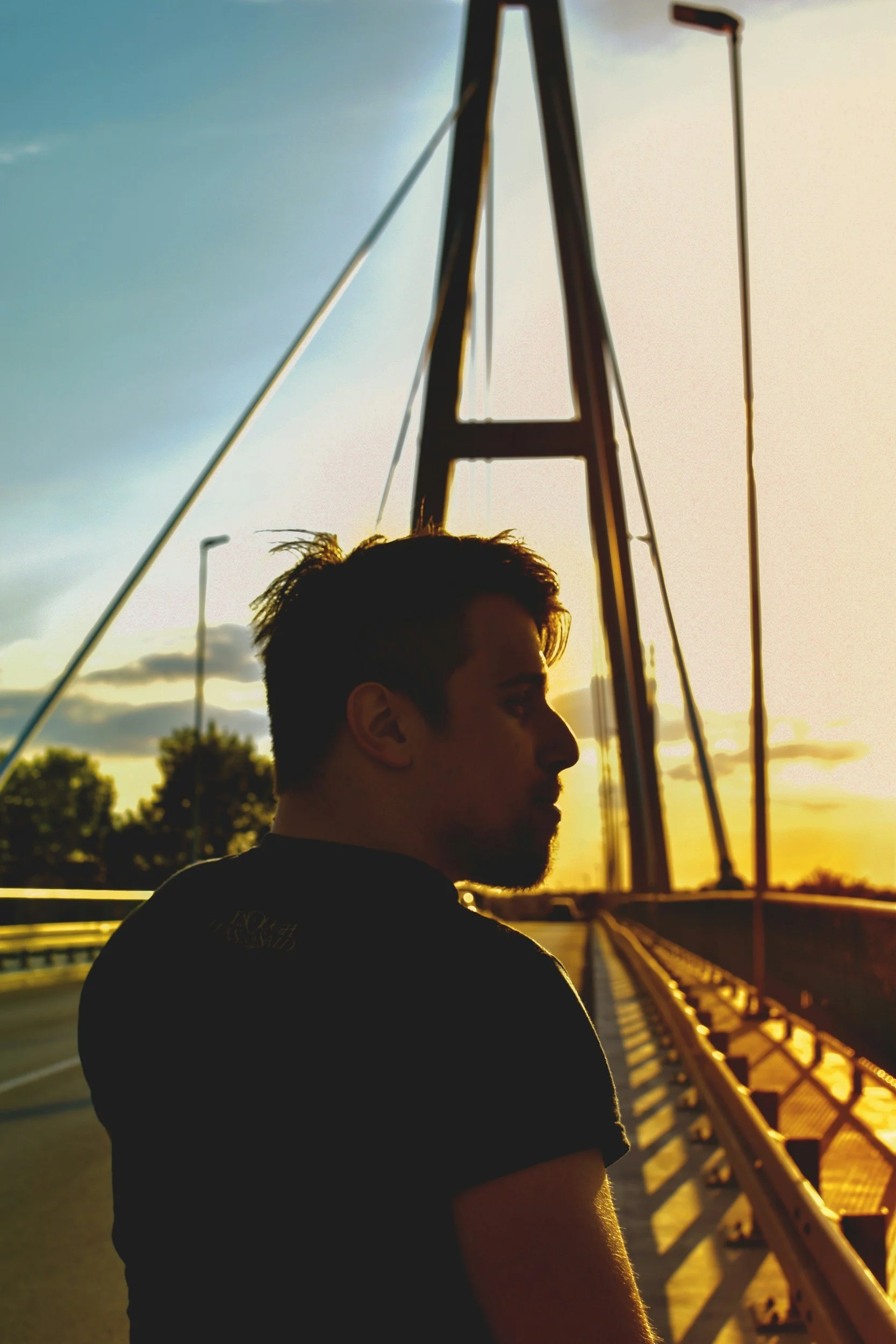 A person with short dark hair and a beard standing on a bridge at sunset, with a large metal structure behind them and a row of streetlights in the distance.