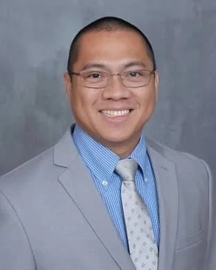 A man in a gray suit, blue dress shirt, and patterned tie, smiling against a plain background.