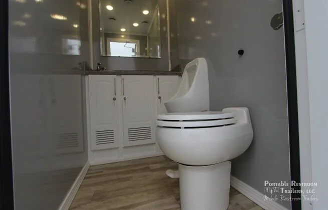 Interior of a portable restroom with a white toilet and a mirror above a sink, wood-look flooring, and white cabinetry.