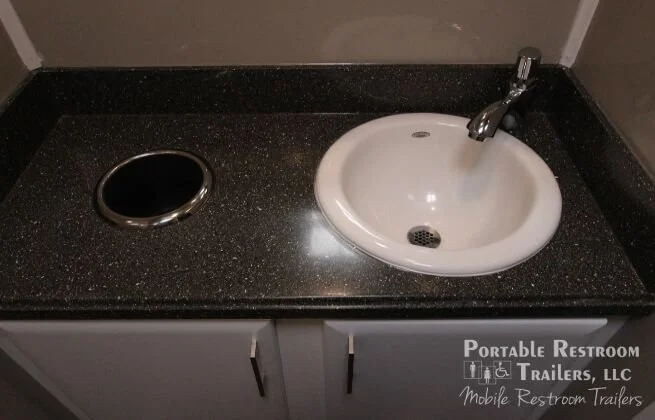 A black countertop with a sink and a separate circular hot plate or drain cover, in a portable restroom trailer.