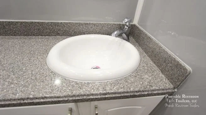 A white sink mounted on a speckled gray countertop with a silver faucet, against a gray wall. Logo in the bottom right corner reads "Portable Restroom Trailers, LLC."
