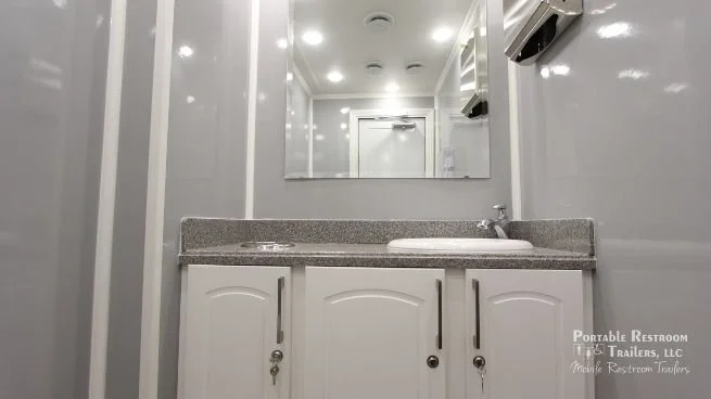 Interior of a portable restroom bathroom with a gray countertop, white sink, and cabinetry, with a mirror on the wall and fluorescent lighting.