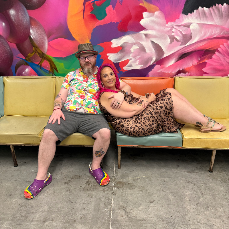 A man and woman sitting on a colorful vintage couch in front of a vibrant, abstract floral mural. The man has a beard, glasses, and is wearing a hat, a bright floral shirt, gray shorts, and colorful Crocs. The woman has purple hair, wearing a leopard