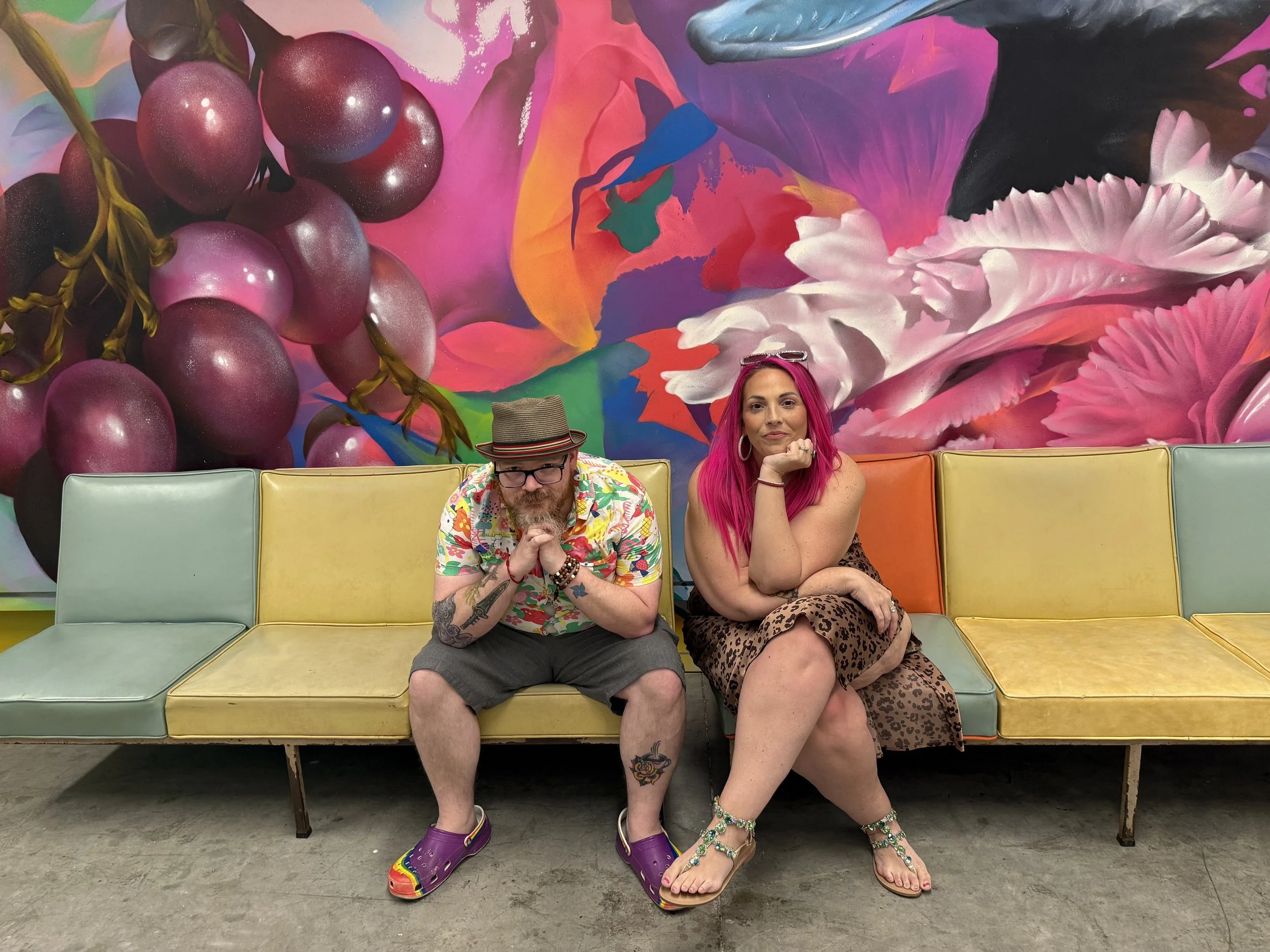 Two people sitting on a vintage, multicolored bench in front of a vibrant, abstract floral mural. The man on the left has a beard, tattoos, glasses, a hat, and colorful clothing. The woman on the right has bright pink hair, large hoop earrings, jewel