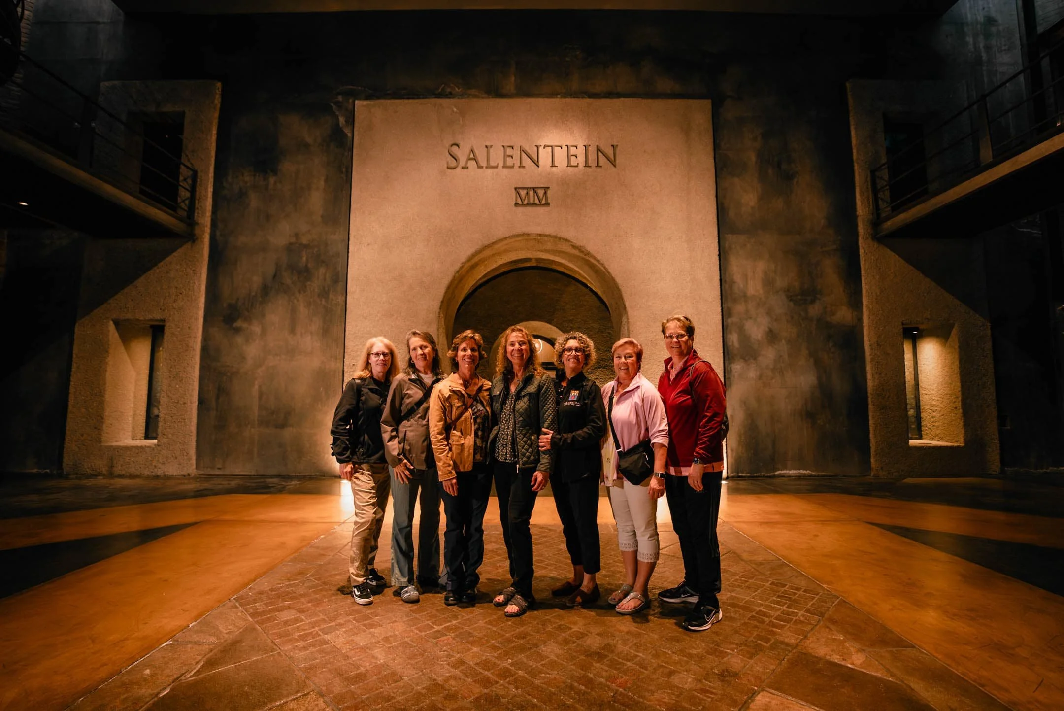 A group of seven women standing together indoors in front of a large wall with the word 'Salentein' and a Roman numeral, with two balconies on each side of the wall.