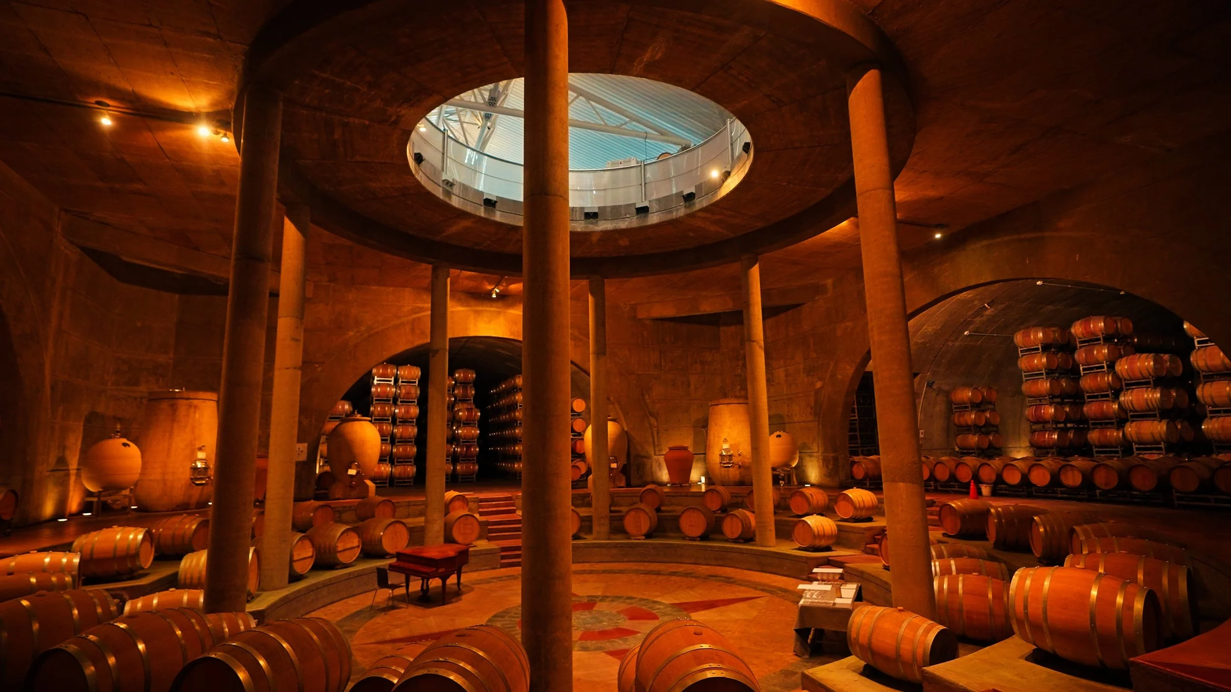 Interior of a wine cellar with stacked barrels on shelves and clay vessels, illuminated with warm lighting, featuring a circular balcony overlooking the space.