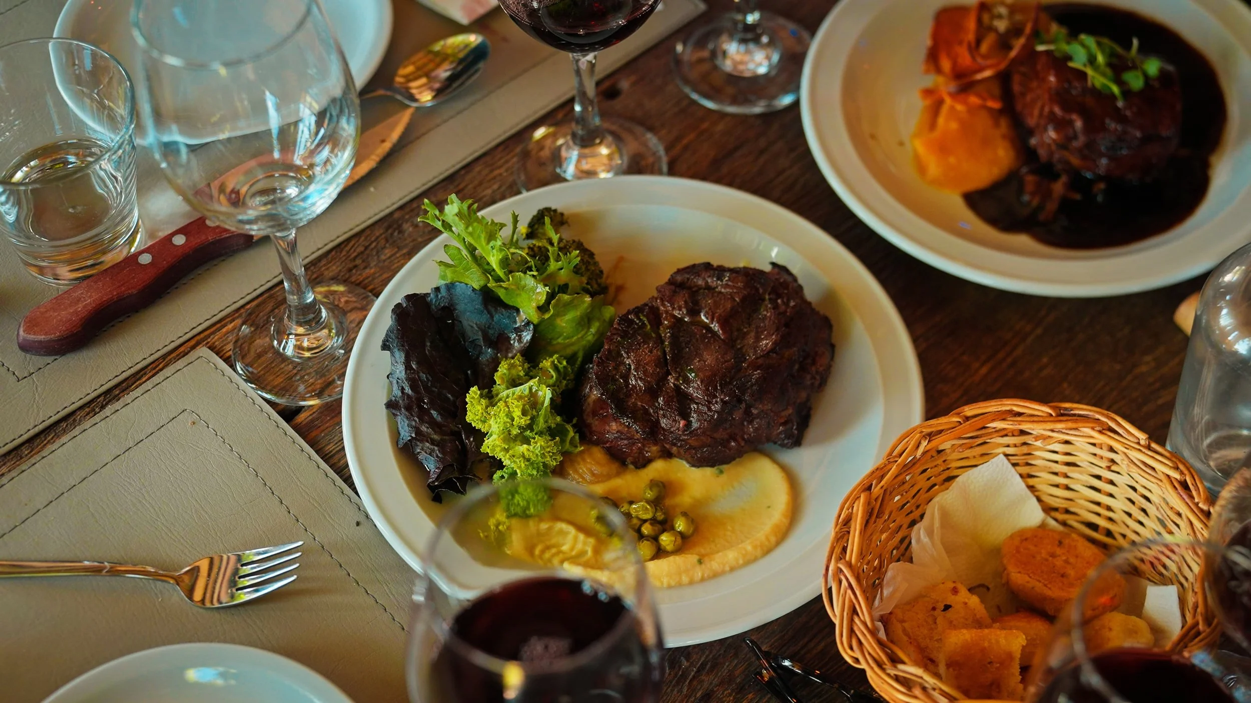 A dinner table with plates of meat, vegetables, and bread, along with glasses of red wine and water.