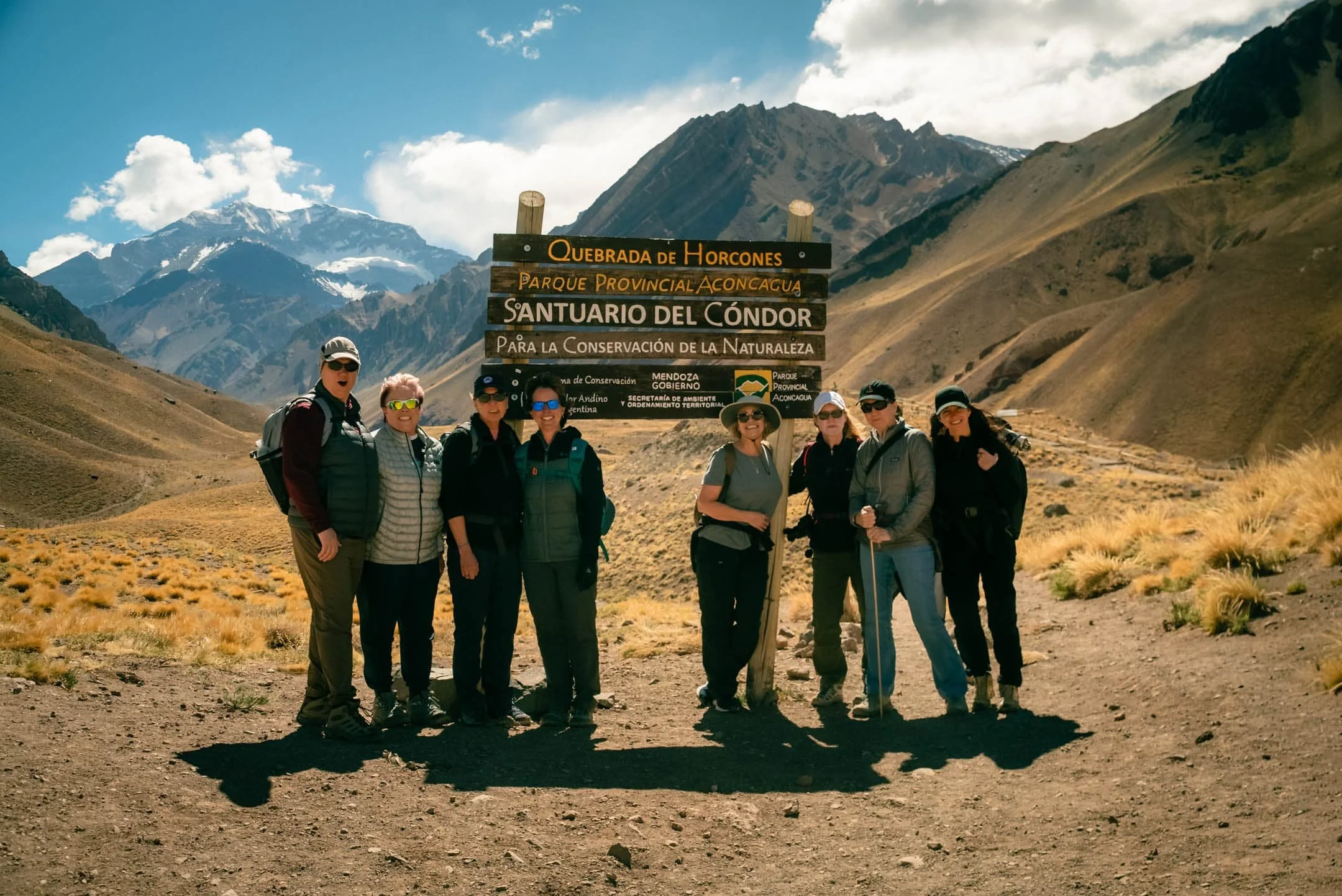 A group of seven hikers standing outdoors in a mountainous area with snow-capped peaks and a partly cloudy sky. They are smiling and wearing hiking gear, including hats, sunglasses, and backpacks, beside a wooden signpost with various location names 