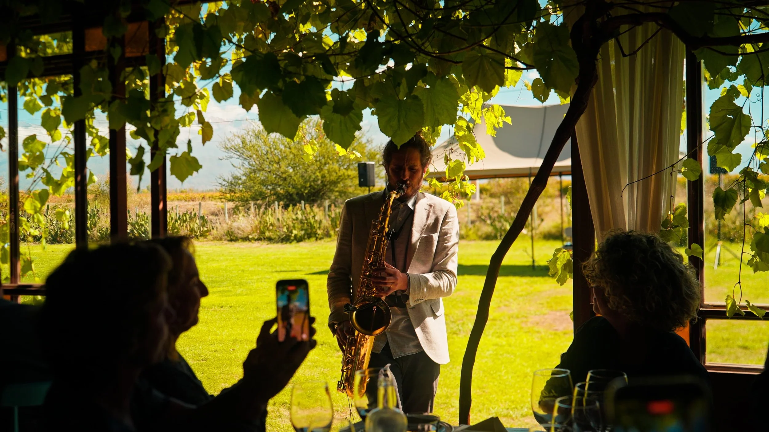 A man in a light-colored blazer playing a saxophone at an outdoor gathering under a shaded trellis with green vines. Guests seated at tables are watching and recording him, with a scenic landscape and blue sky in the background.