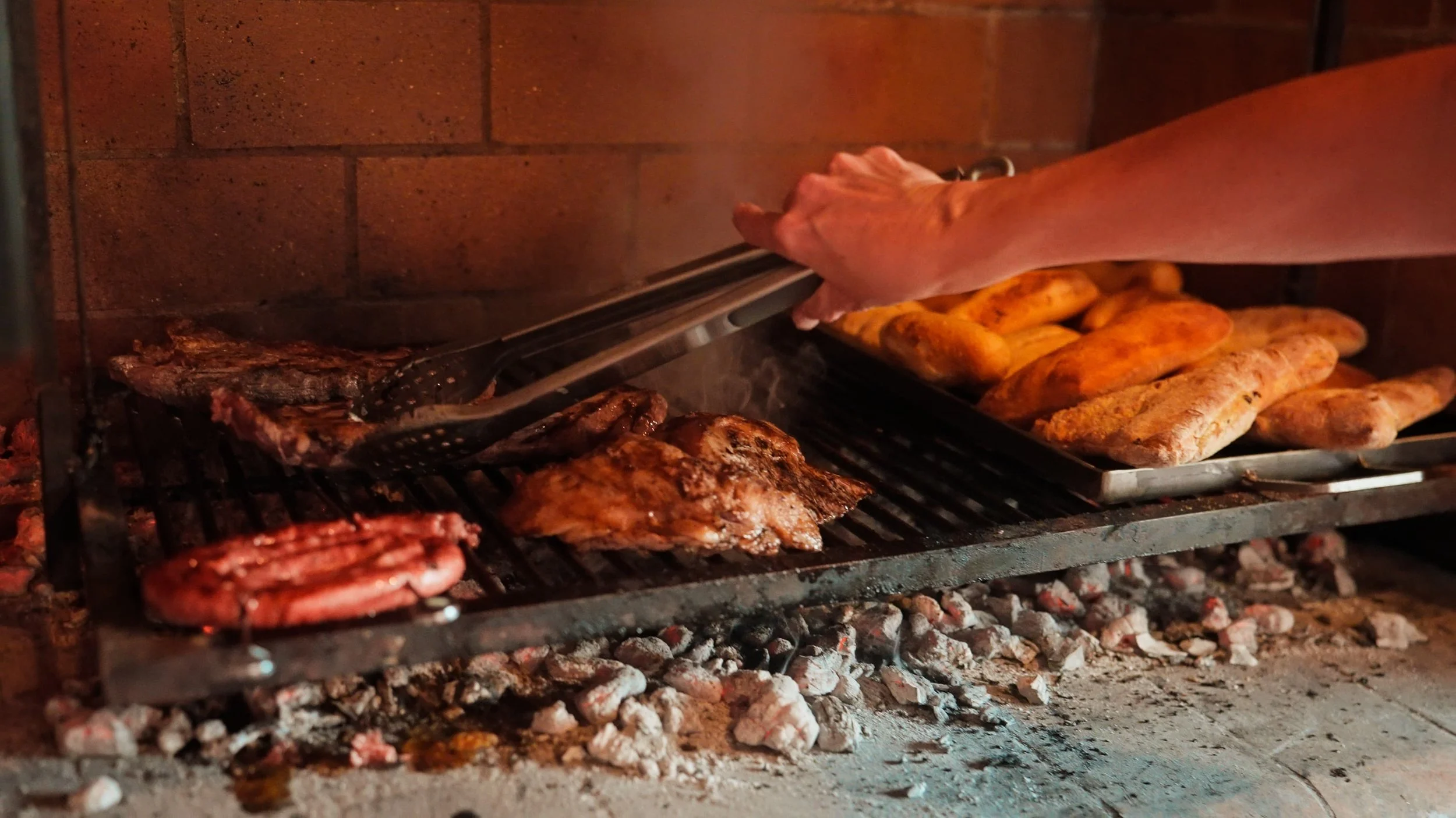 A person placing baked bread on a grill inside an oven, with a background of orange bricks and burning coals below.