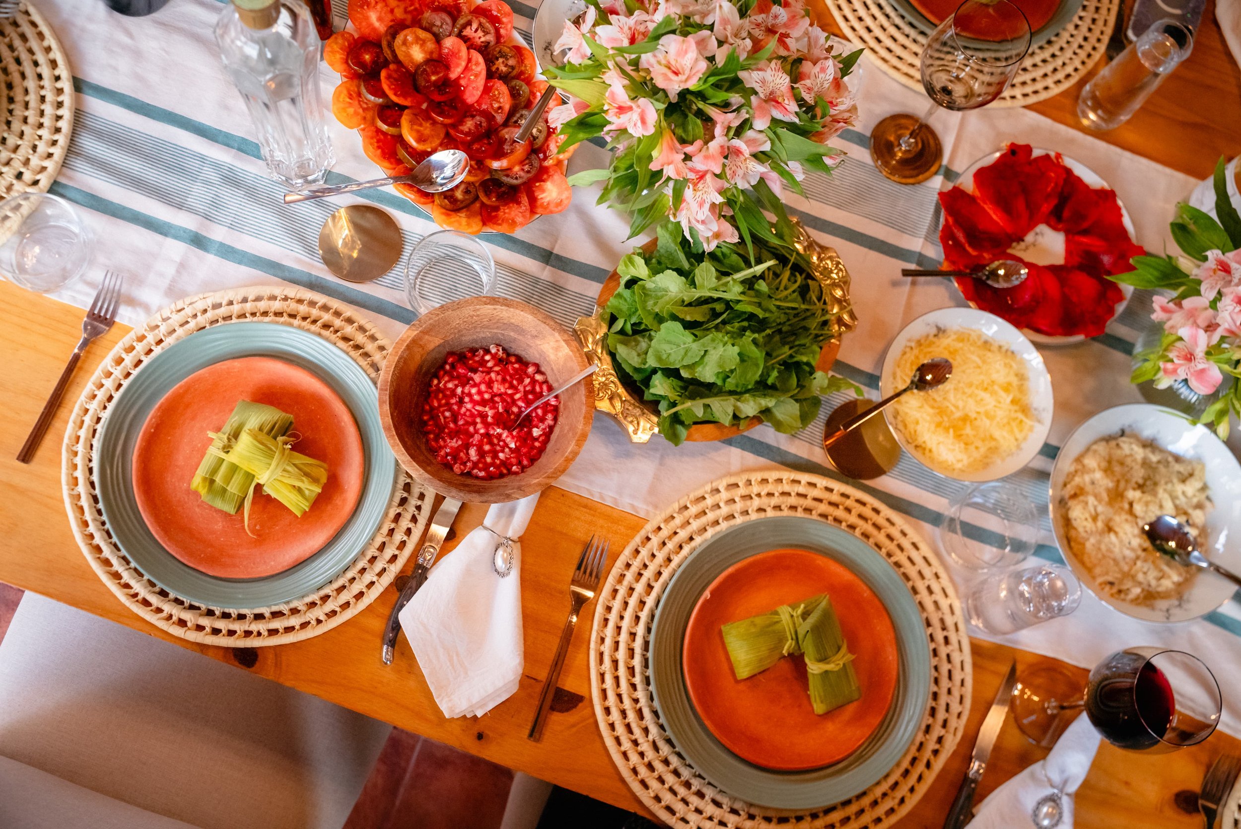 A dining table set with plates of tomato soup garnished with celery, bowls of pomegranate seeds, cheese, and other dishes, with floral centerpieces and glassware.