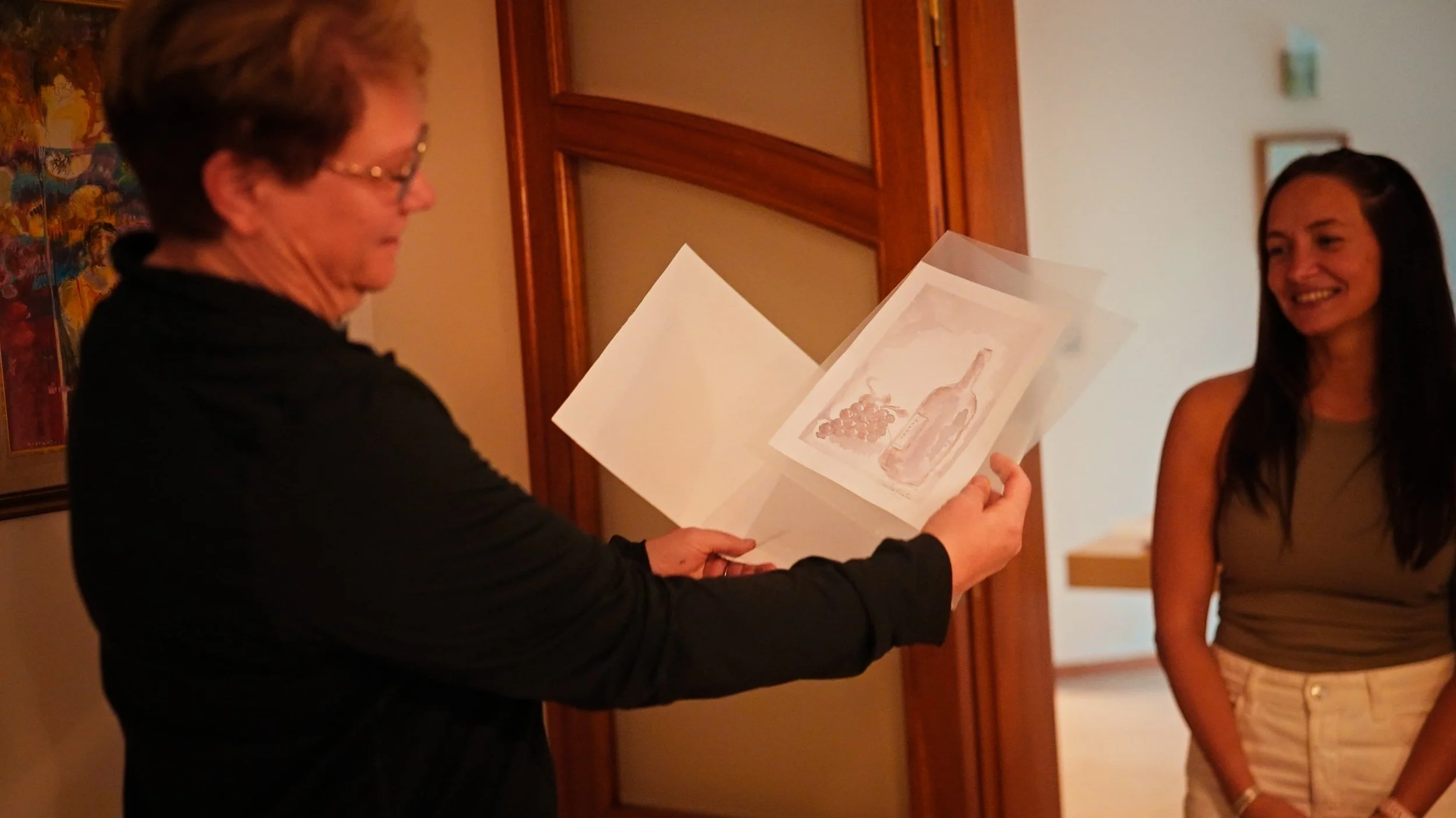 An older woman is giving a gift of a watercolor illustration to a young woman, who is smiling. The watercolor features a bottle of wine, a bunch of grapes, and a wine glass. They are indoors, near a wooden door and some colorful artwork on the wall.
