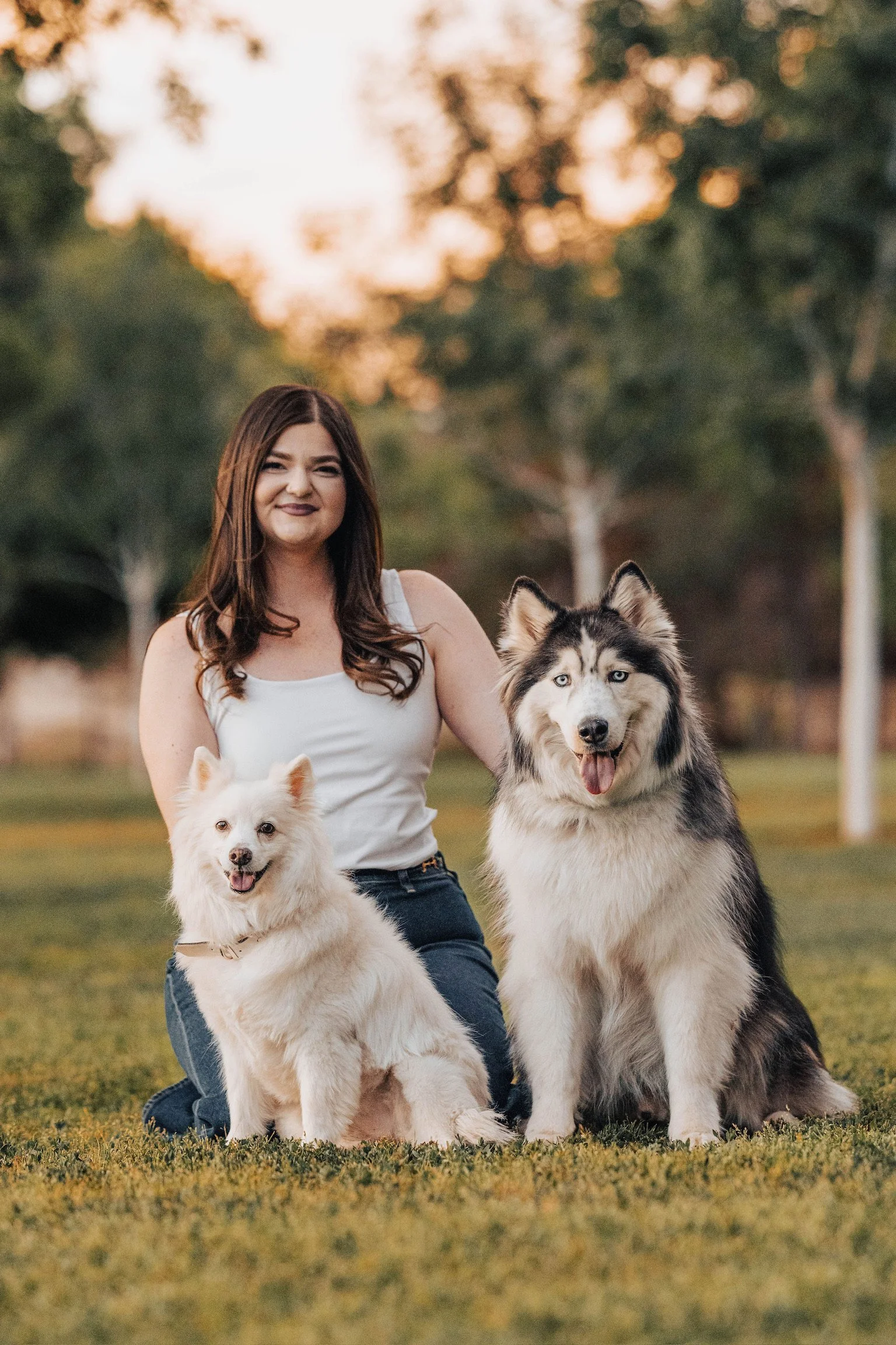 A woman with brown hair sitting with two Siberian Huskies on a grassy field during sunset, with trees in the background.
