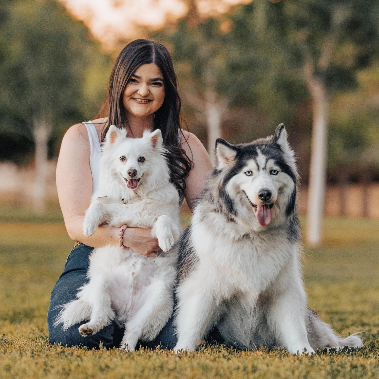 A woman smiling and squinting while holding a white dog with fluffy fur, and sitting next to a large Siberian Husky with blue eyes, on a grassy area outdoors with trees in the background during sunset.