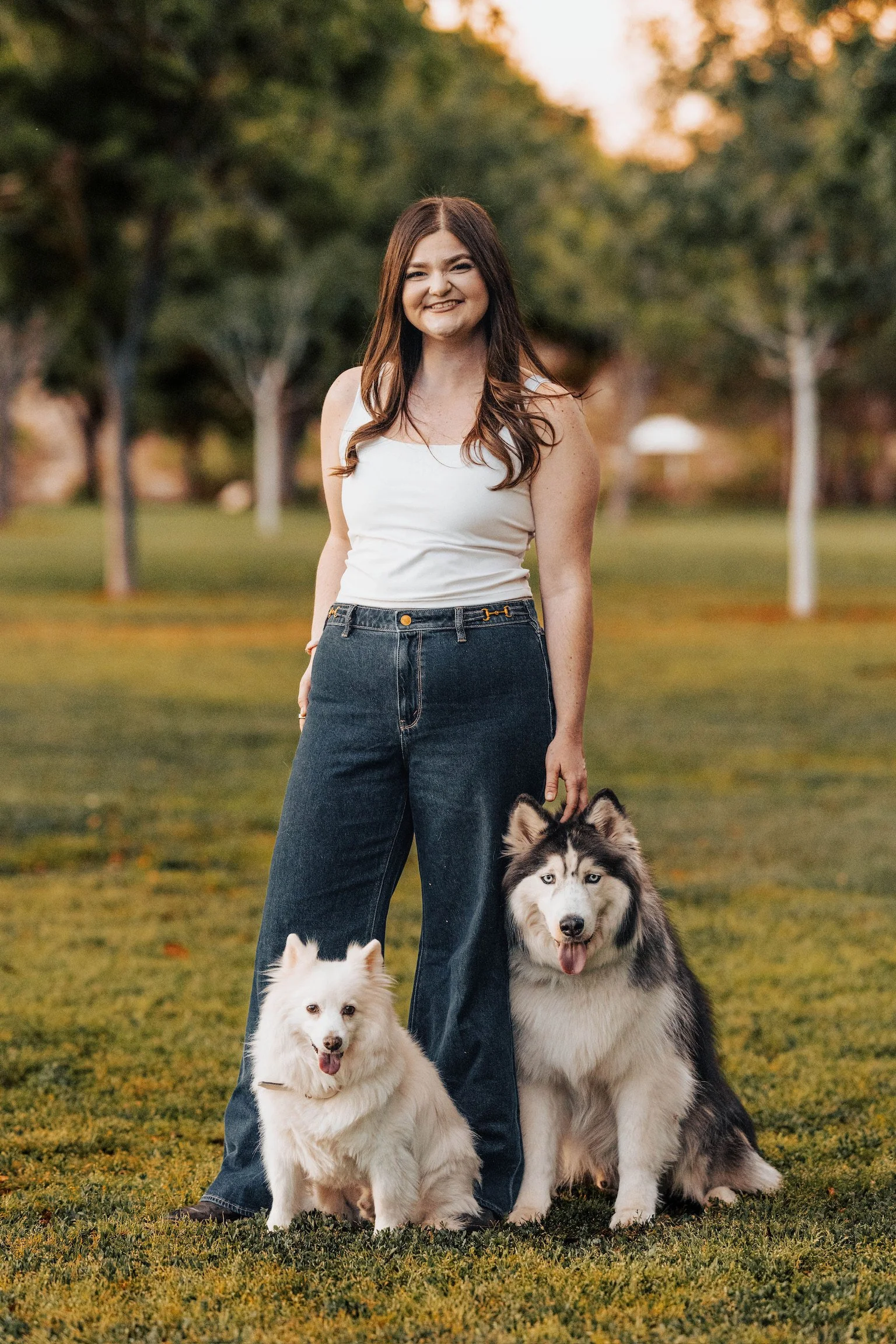 Smiling young woman in white tank top and jeans standing outdoors with two dogs, a fluffy white dog and a large Siberian Husky, on grassy field with trees and sunset in the background.