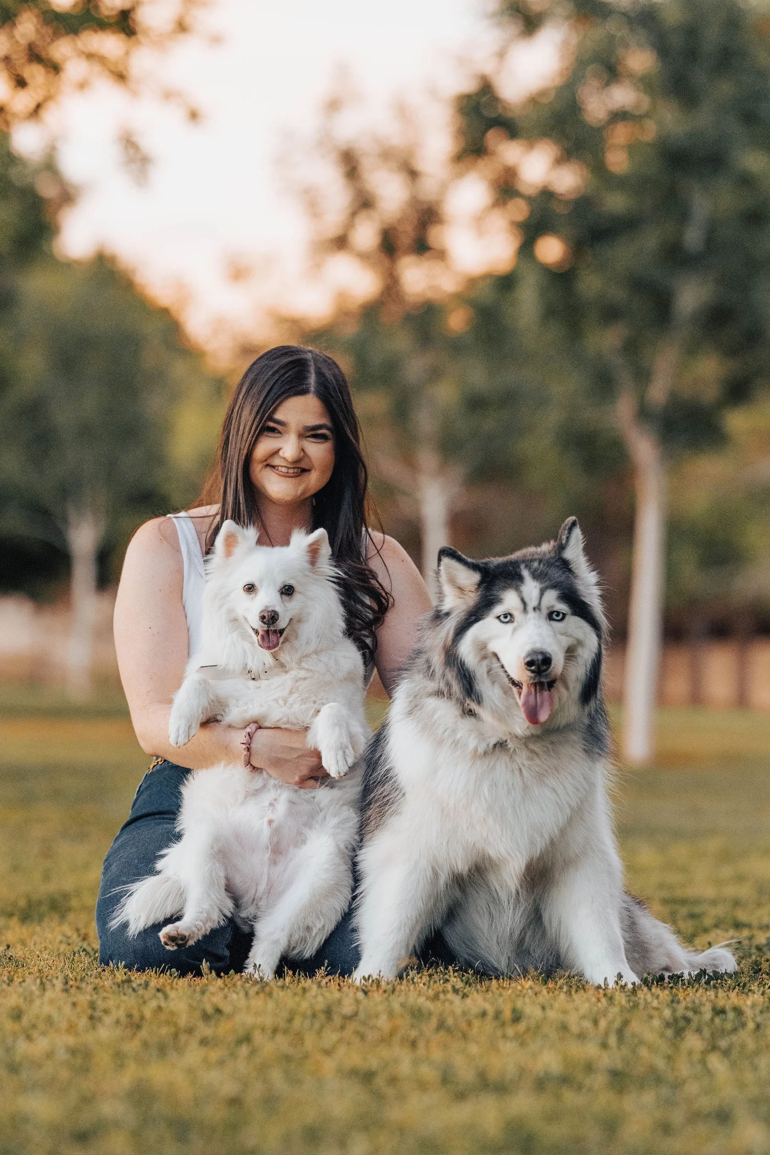 A woman smiling and holding a white dog, with a large Siberian Husky sitting beside her in a park at sunset.