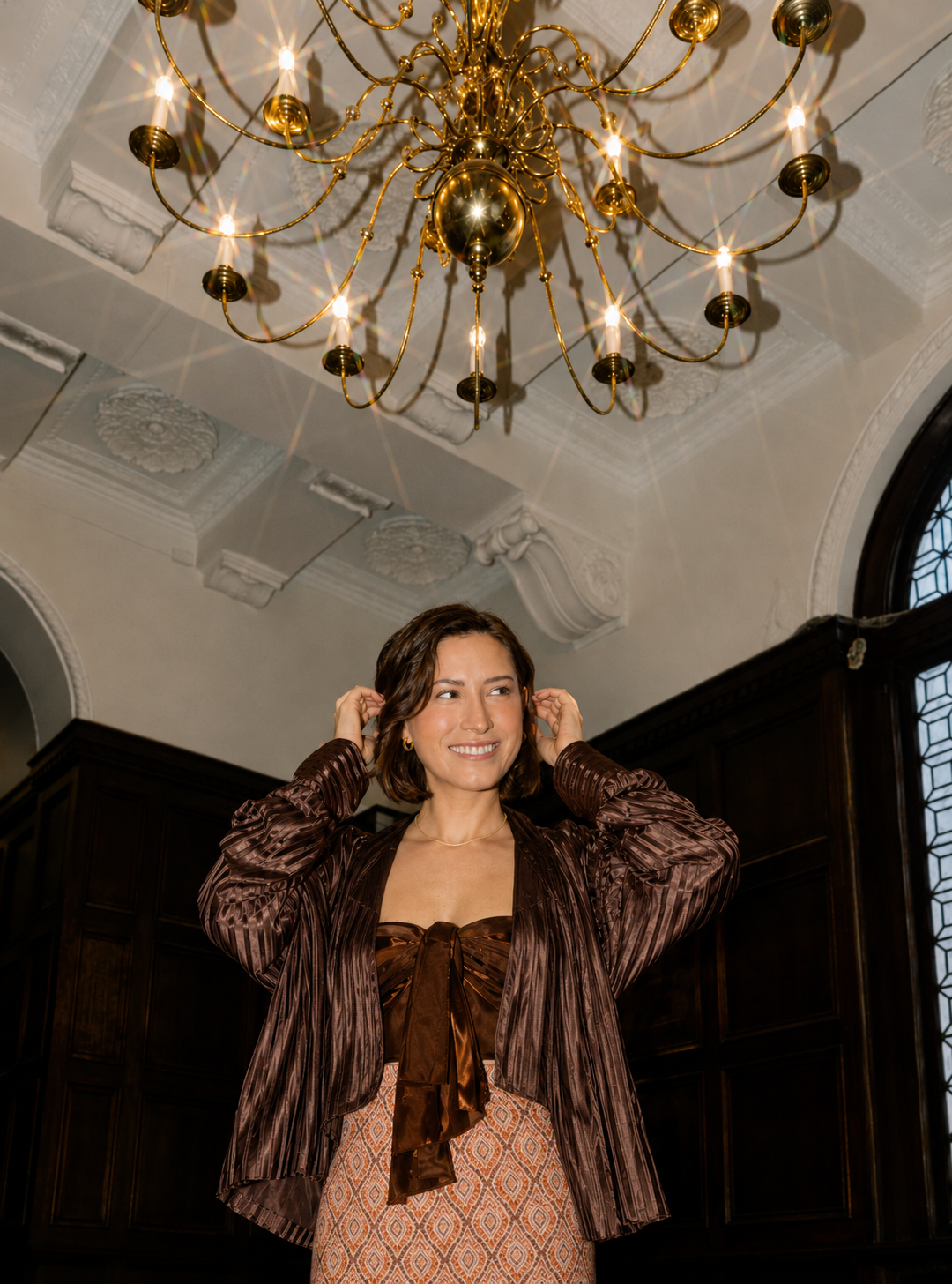 A woman smiles and adjusts her hair in a room with a grand chandelier and dark wood paneling.