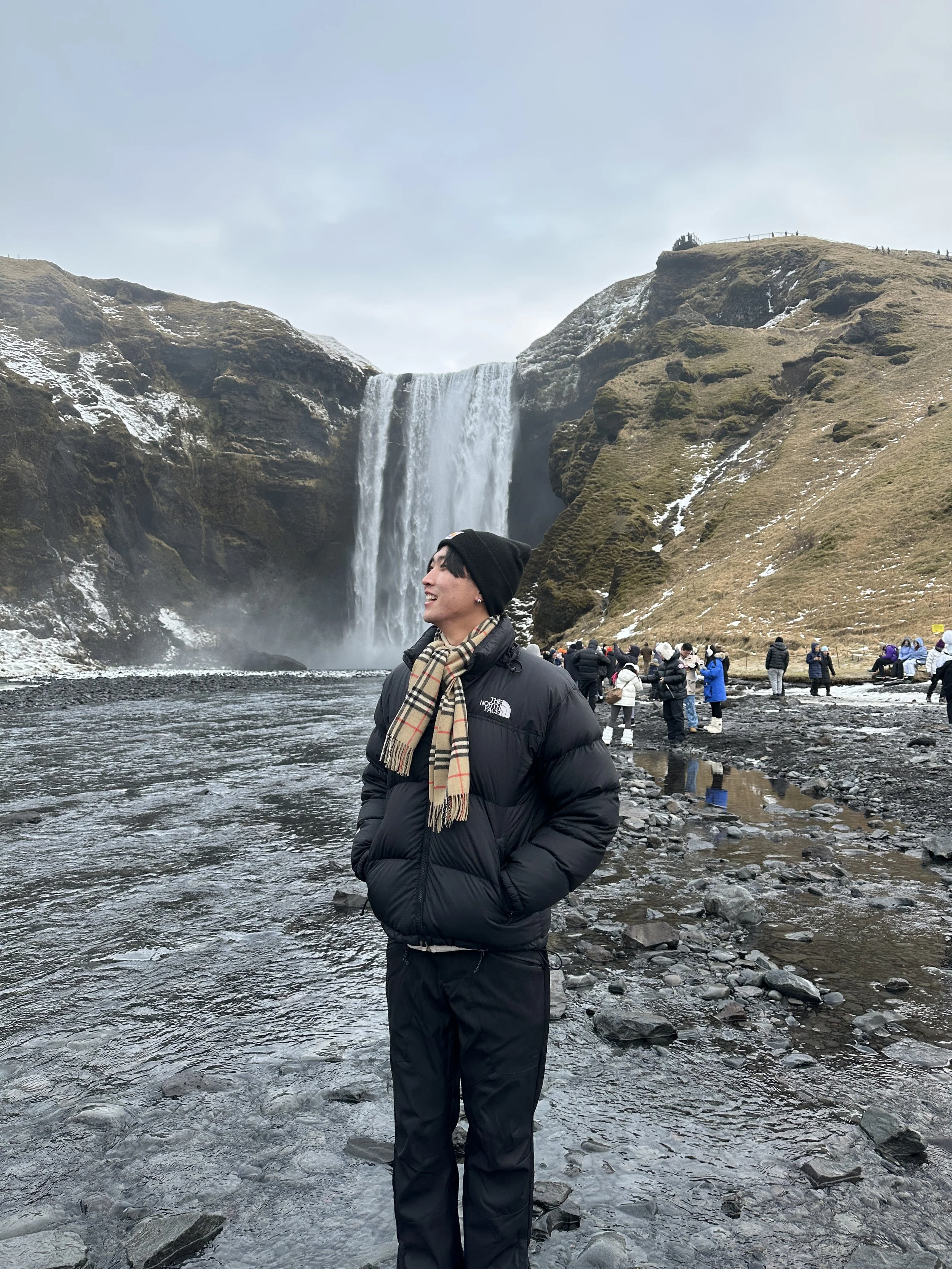 A person standing by a river with a waterfall and mountain landscape in the background, people in winter clothing on the rocky shore.