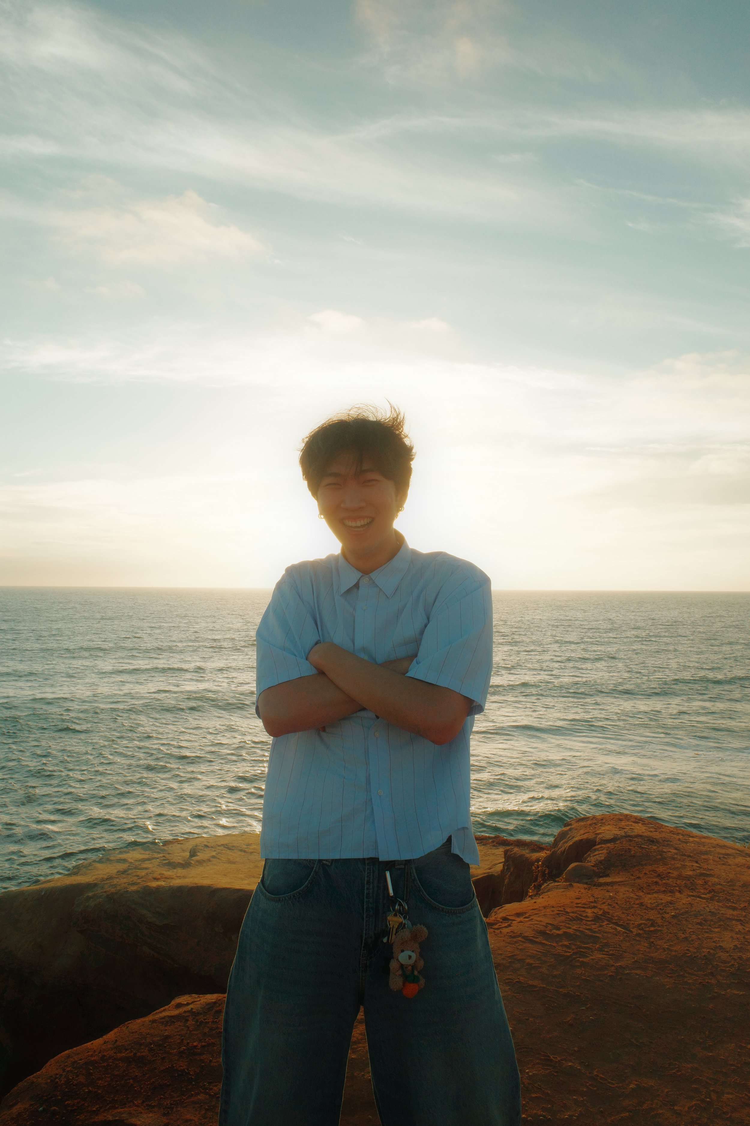 A person with short dark hair smiling and crossing their arms in front of the ocean at sunset, with rocks visible at the bottom of the image.