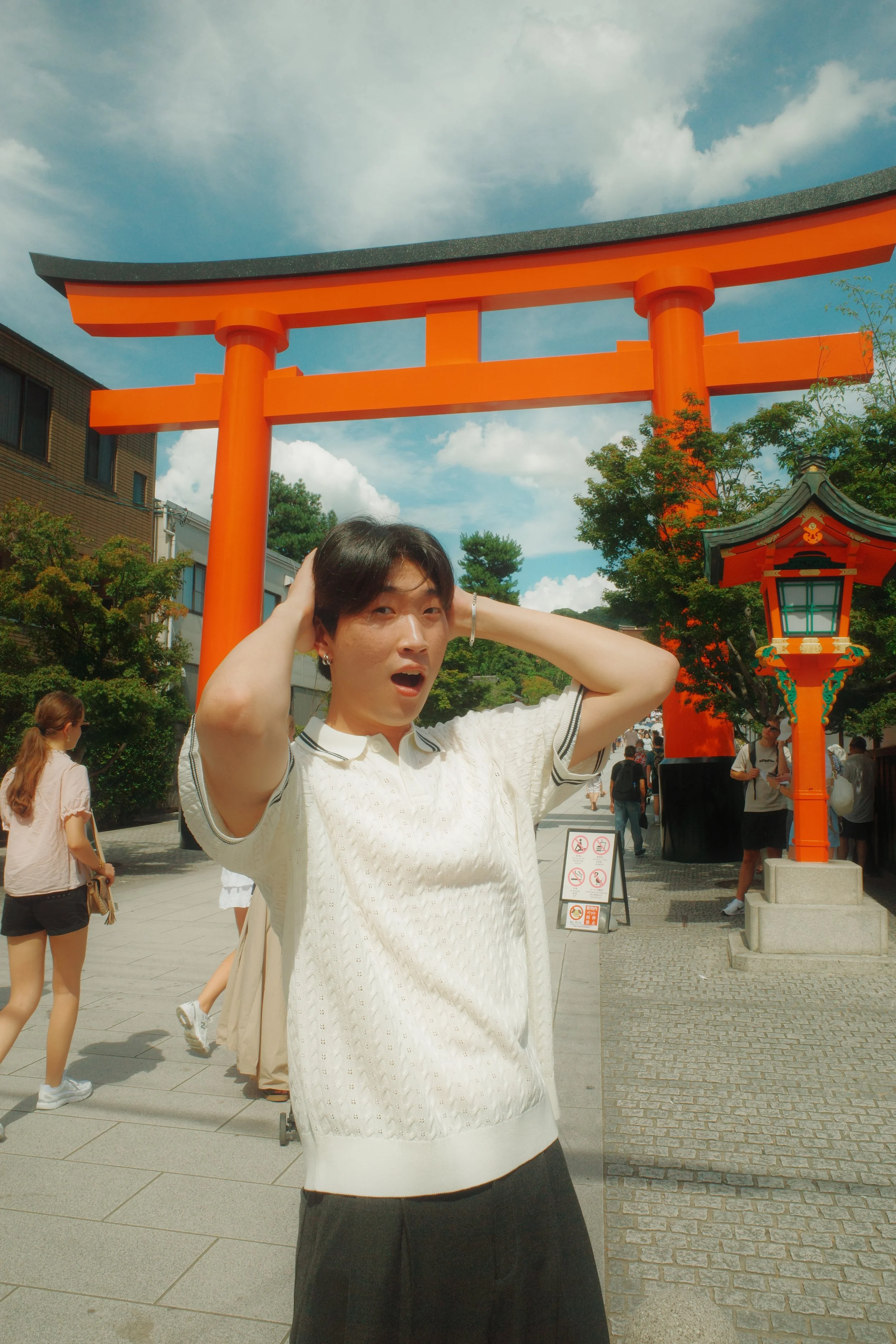 A person standing in front of a bright red Torii gate, typical of Japanese shrines, with a blue sky and some clouds overhead. The person has short dark hair, is wearing a white shirt with black trim, and appears to be surprised or amused.
