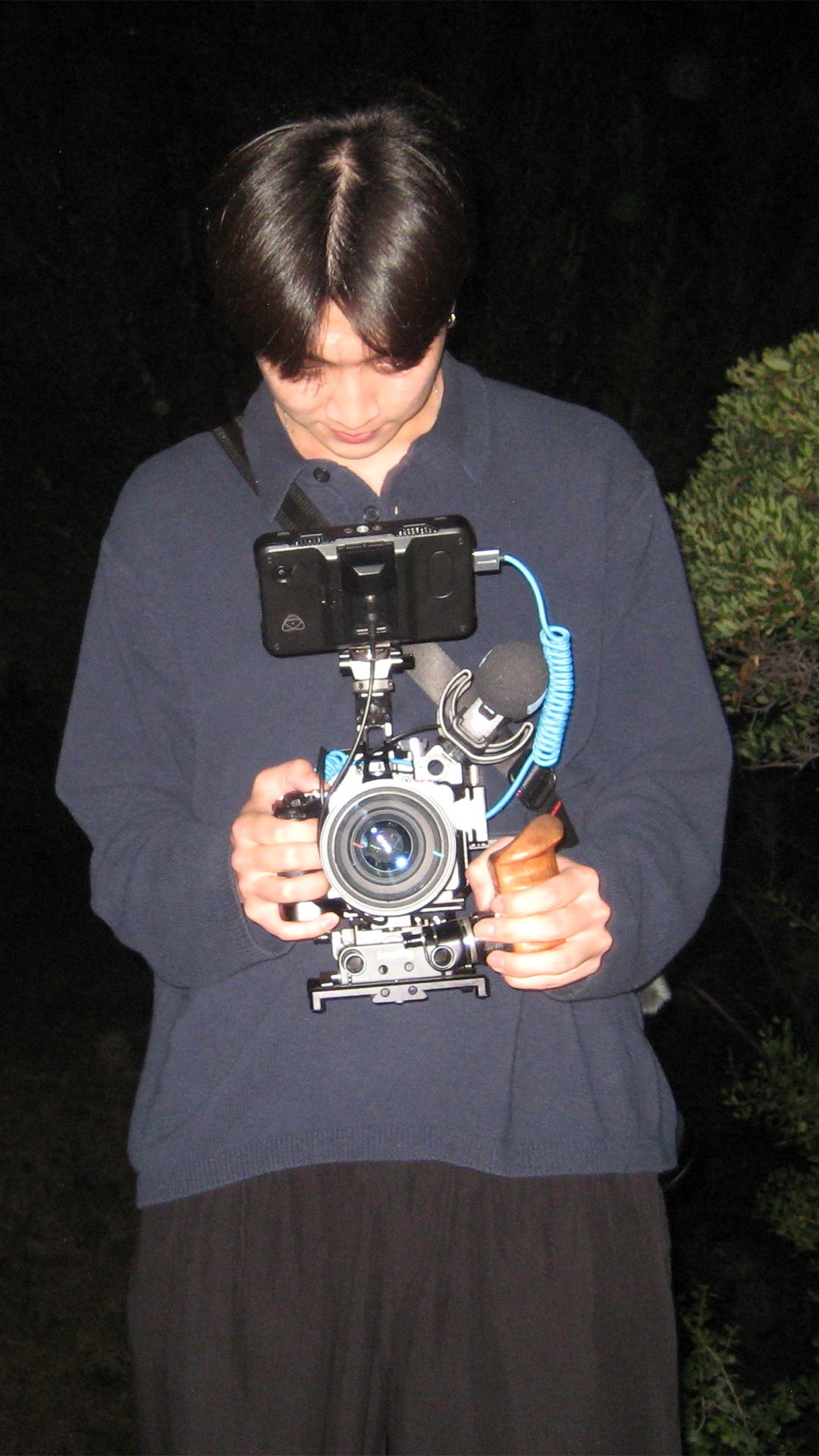 A person with short dark hair holding a professional camera setup with a smartphone attached on top, against a dark outdoor background.