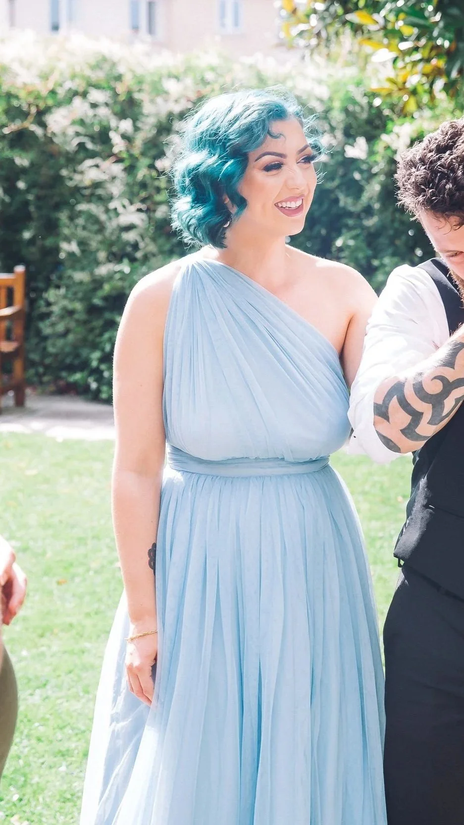 A woman with blue curly hair wearing a light blue, sleeveless, pleated gown, smiling outdoors in sunlight with green bushes in the background.