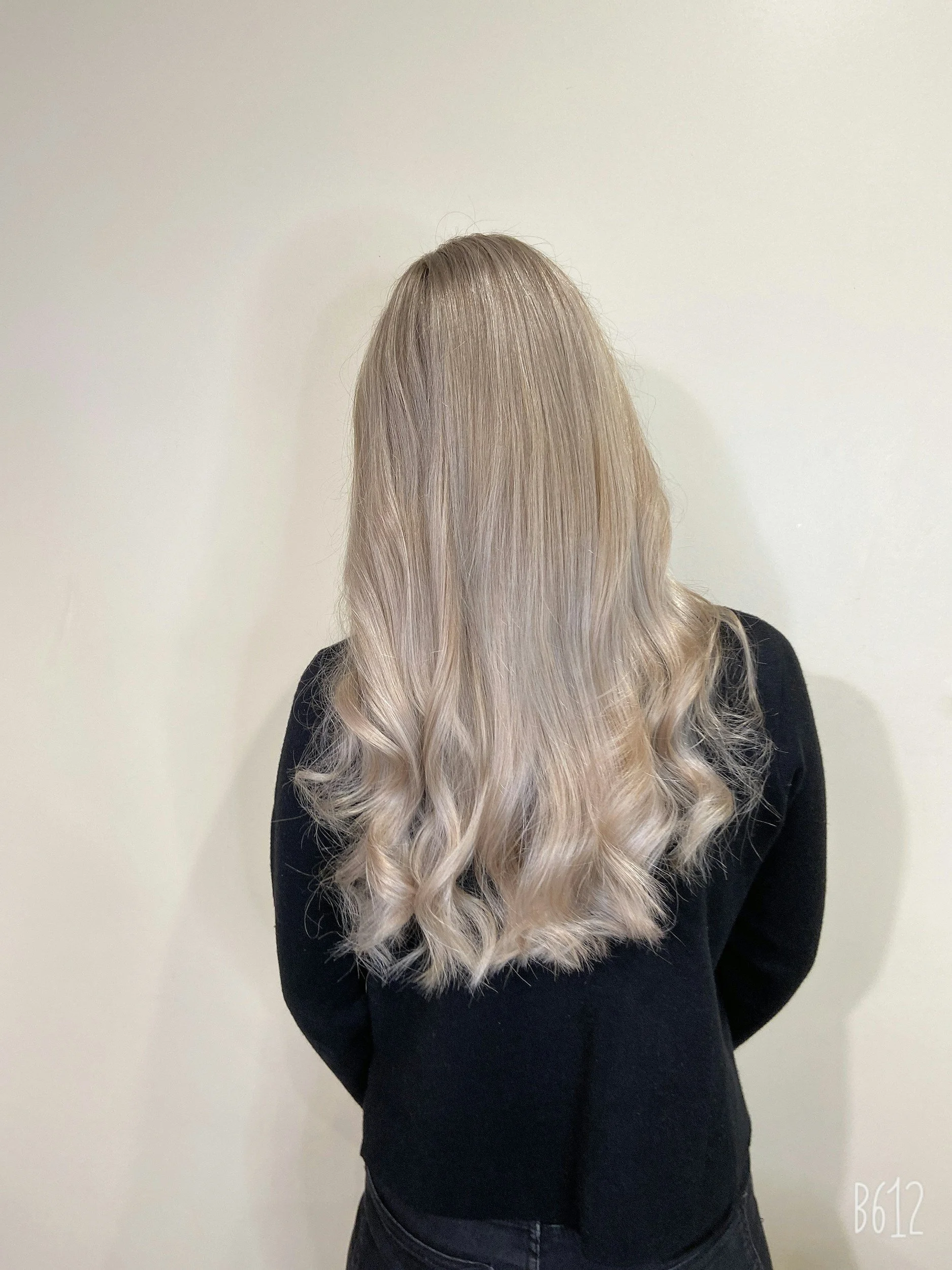 A person with long, wavy, blonde hair standing in front of a plain white wall, wearing a black top and dark jeans.