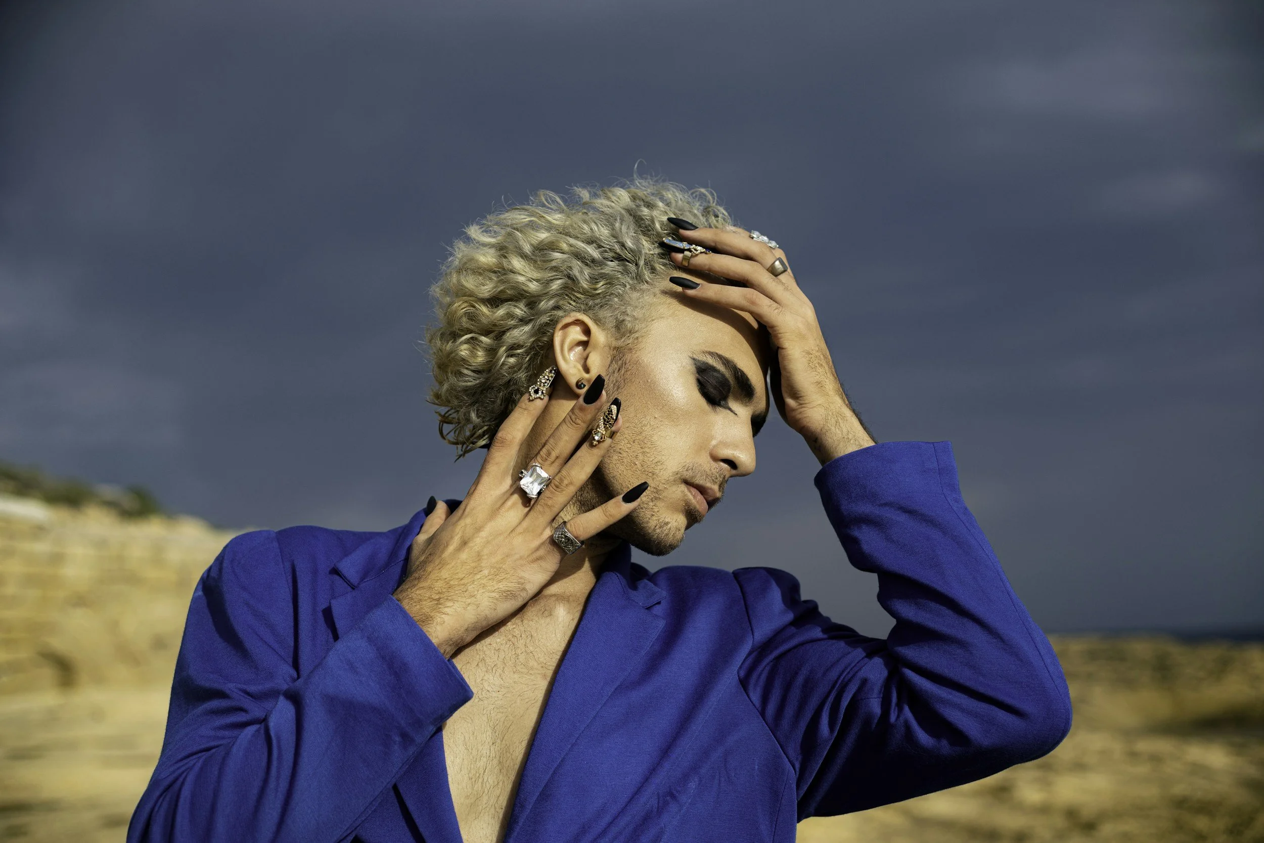 A person with platinum blonde, curly hair and various rings and earrings, wearing a blue shirt, has one hand on their forehead and the other near their face, with a cloudy sky in the background.