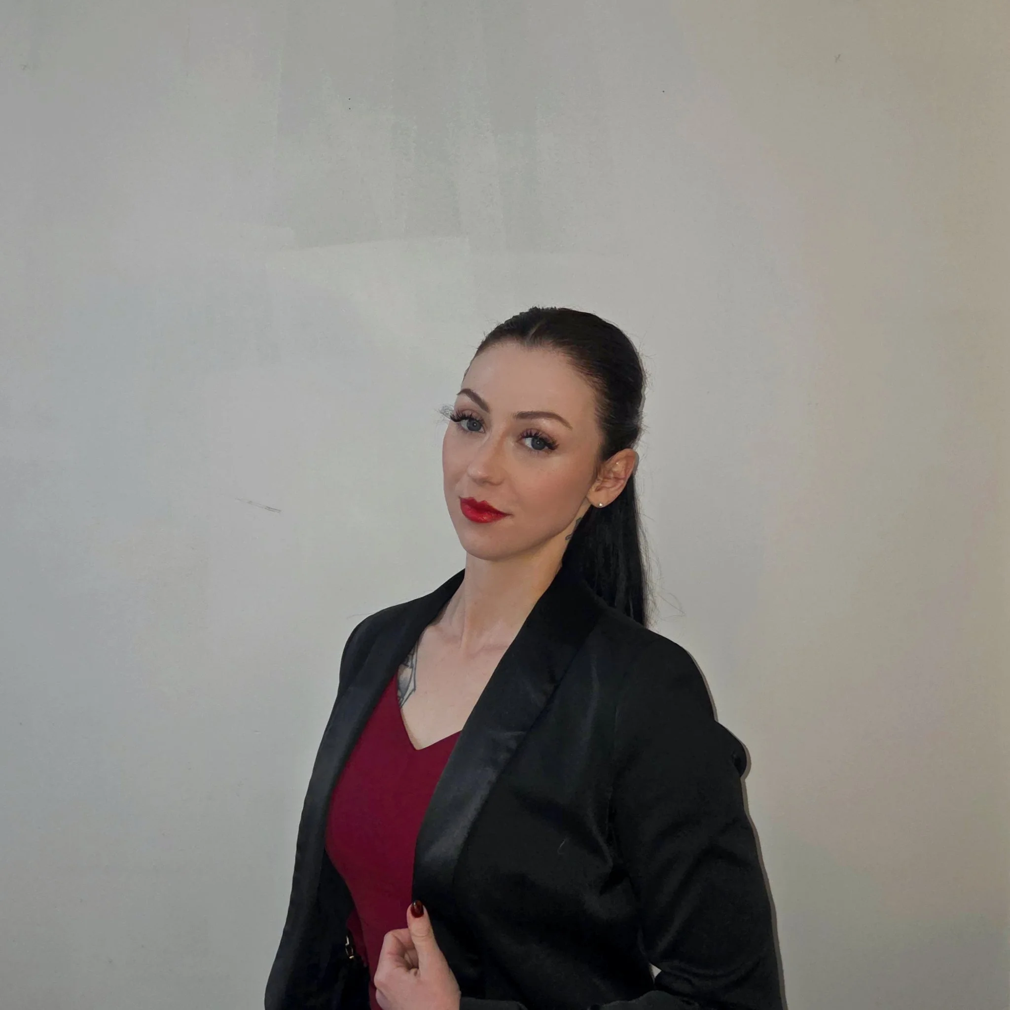 A woman with dark hair tied back, wearing bright red lipstick and a black jacket, is giving a thumbs up while pointing to her chest with her index finger. The background is a plain, light-colored wall.