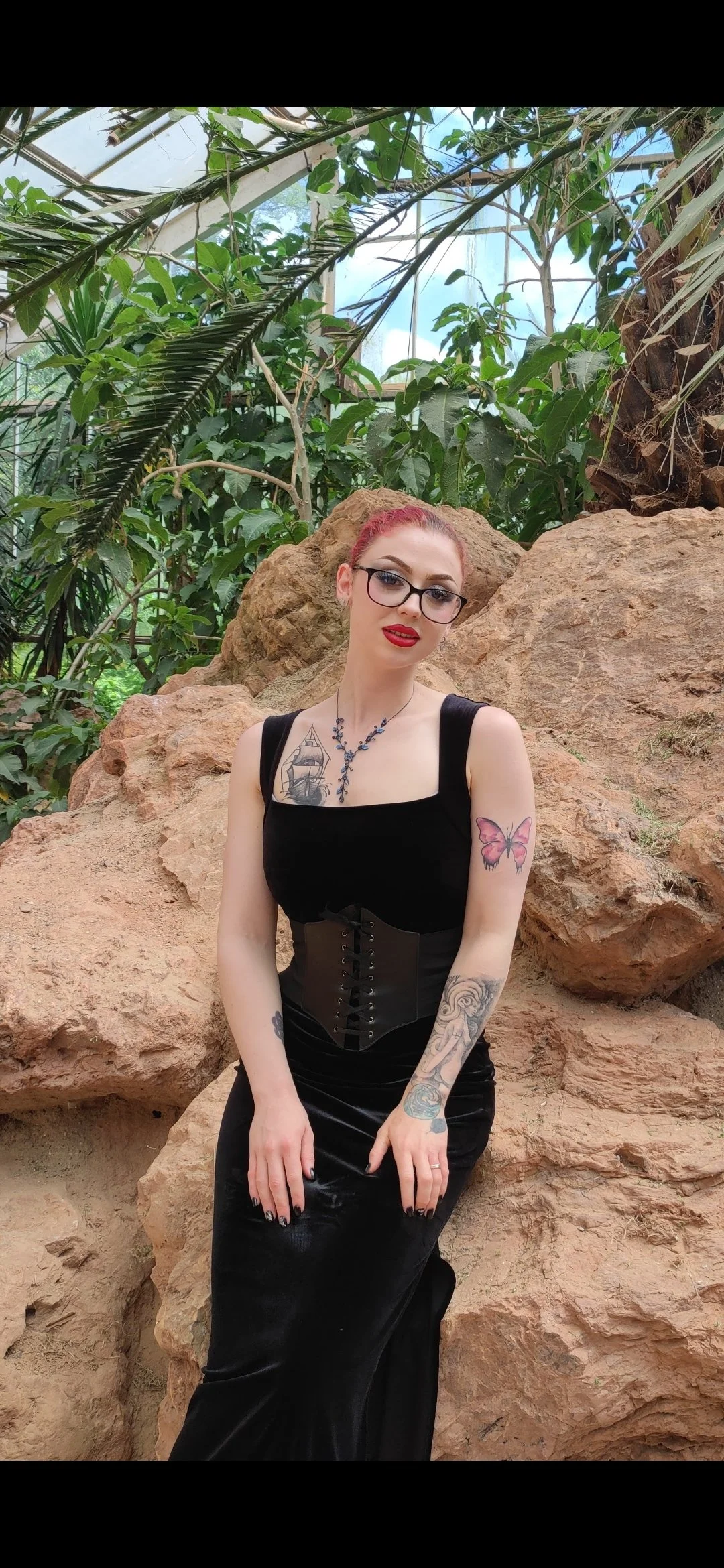 A woman with red hair, glasses, and red lipstick poses in front of rocks and tropical plants in a greenhouse, wearing a black dress with tattoos on her arms and a necklace.
