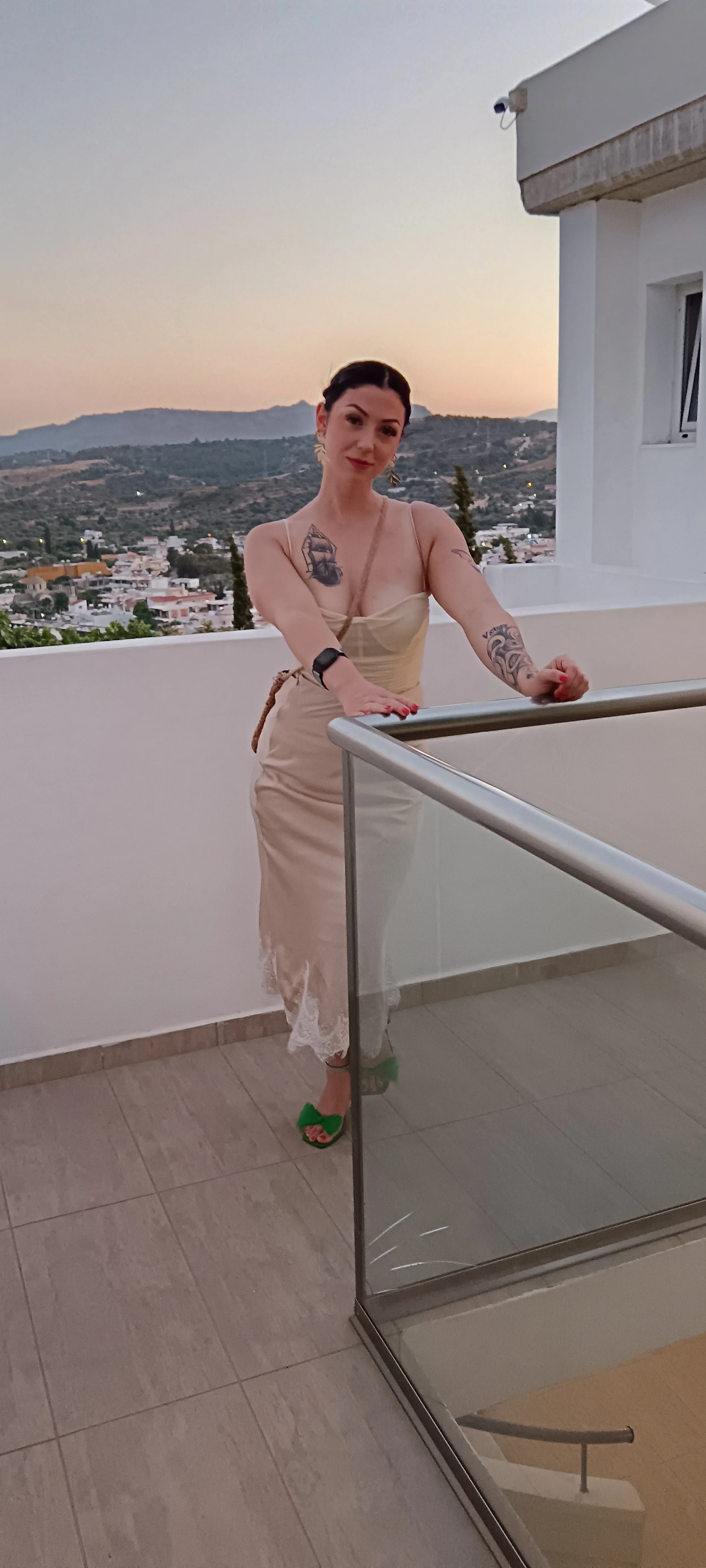 A woman standing on a balcony during sunset, wearing a beige dress and green high heels, with tattoos on her arms and a watch on her wrist, overlooking a town and mountains in the distance.
