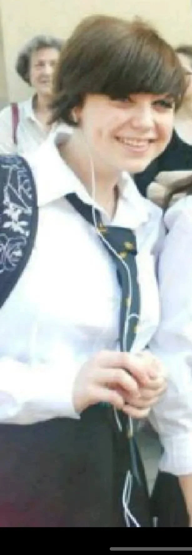 Smiling young woman in a school uniform with a white shirt and dark tie, carrying a backpack, standing indoors.