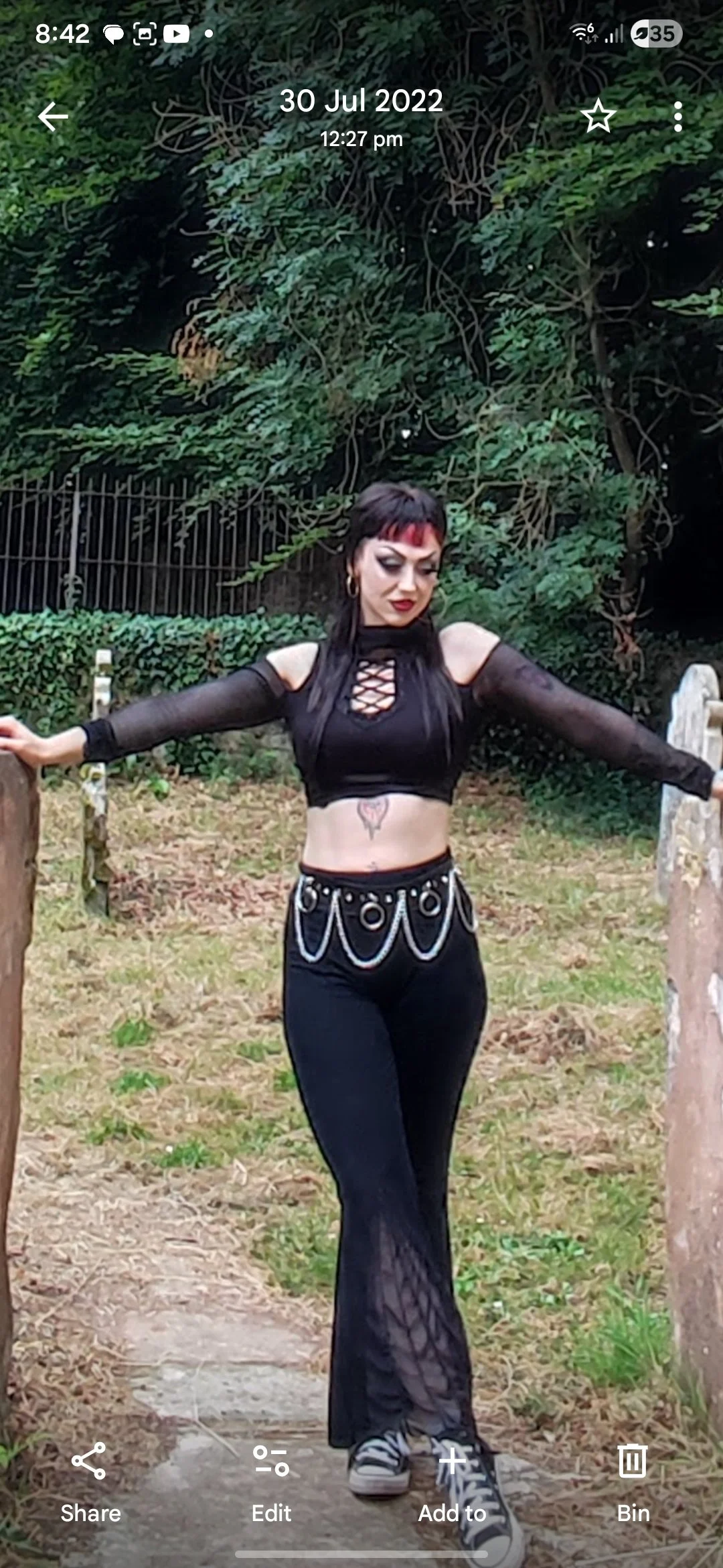 A woman with black hair and red streaks, wearing gothic-style makeup, a black lace-up crop top with sheer long sleeves, and black mesh pants with a chain belt, standing outdoors with arms outstretched between two stone pillars.