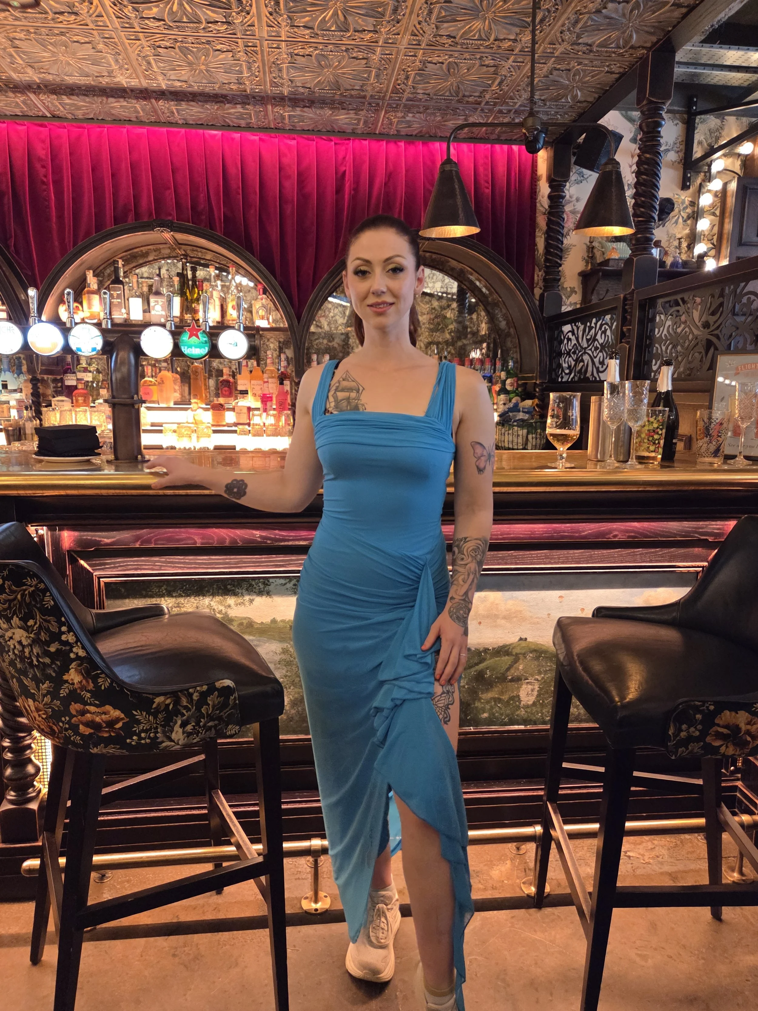 A young woman wearing a blue dress and white sneakers standing at a bar with bottles of alcohol behind her, in a dimly lit, vintage-style bar with red velvet curtains and ornate decor.