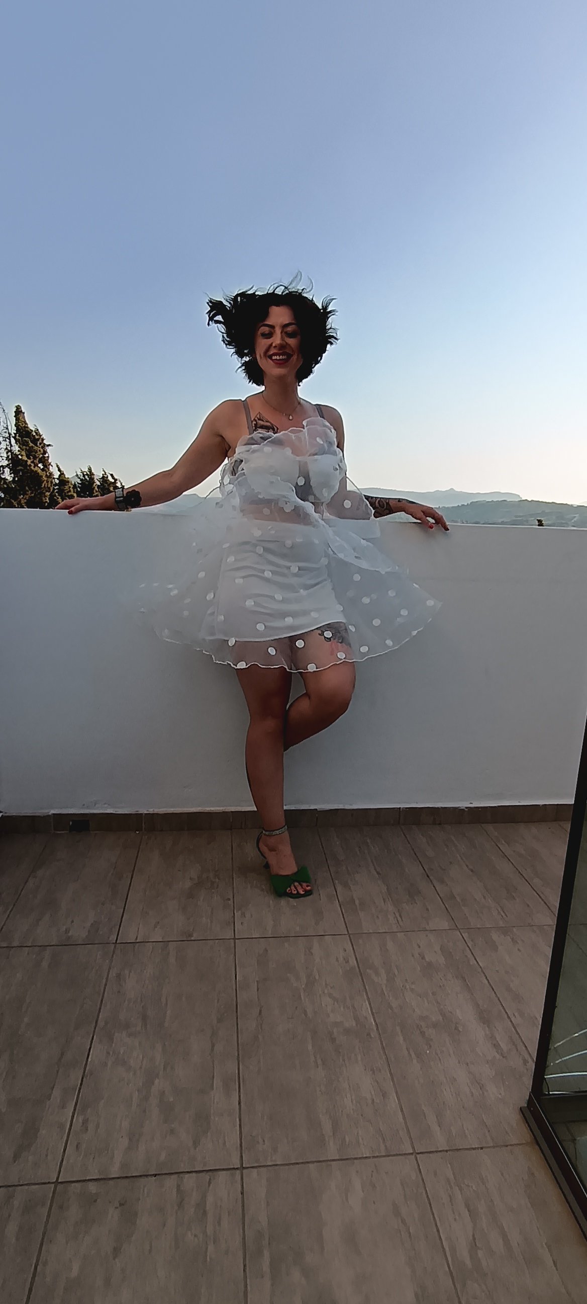 Woman with curly dark hair wearing a sheer, polka-dotted dress and green high heels, standing outdoors against a white wall with a view of hills and trees in the background, smiling.