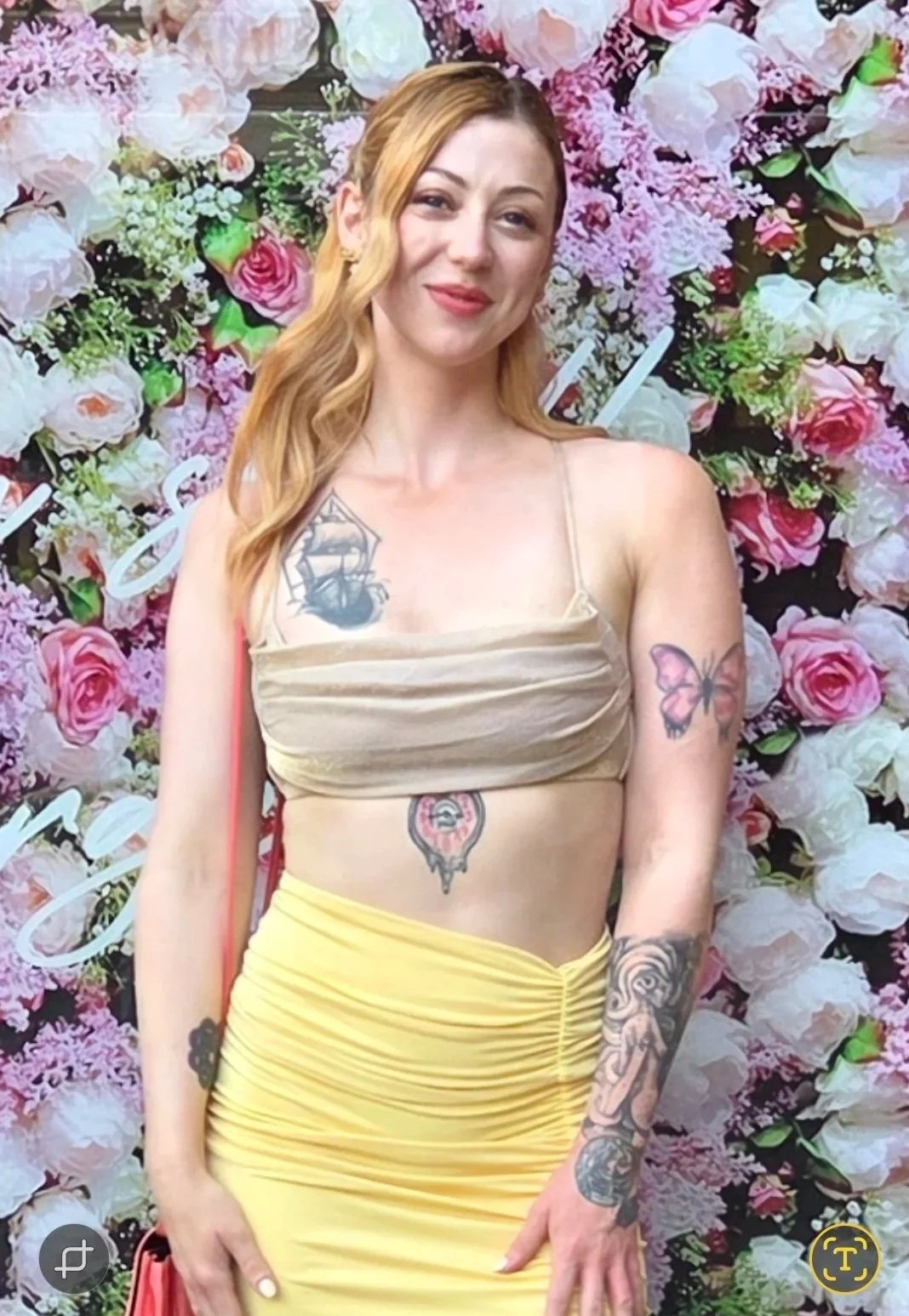 A woman with long wavy red hair poses in front of a flower wall with pink and white roses. She is wearing a beige spaghetti strap top and a bright yellow skirt. She has tattoos on her left arm, right shoulder, and stomach, including a butterfly, a ship, and a clock.