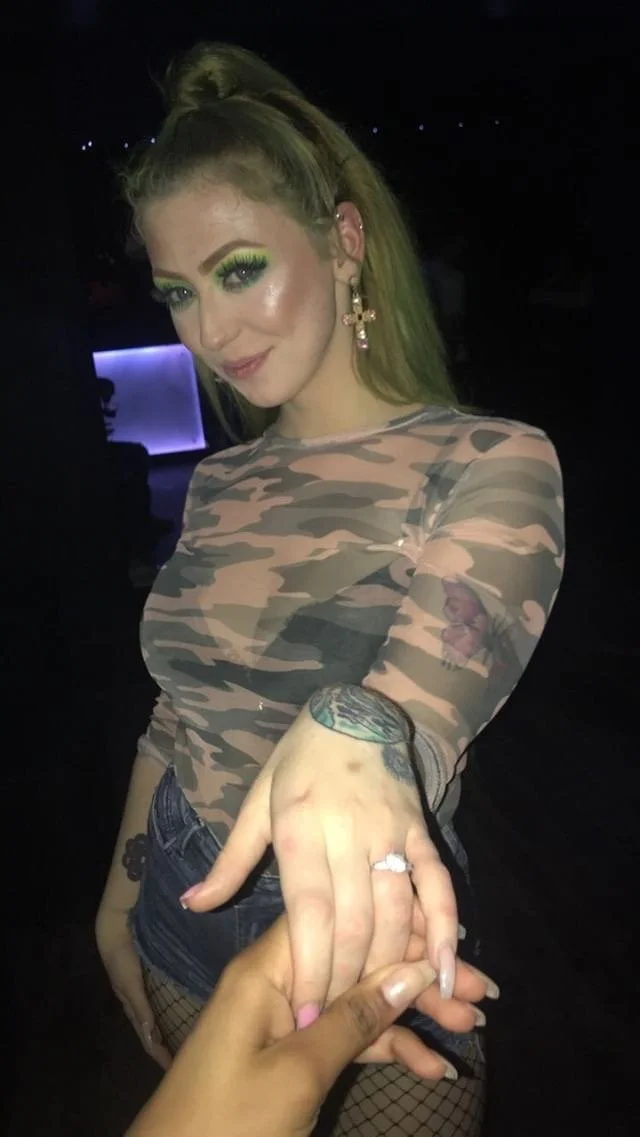 A young woman with green eye makeup and earrings is showing off her hand with a tattoo while holding hands with another person with darker skin. She is wearing a camouflage-patterned long sleeve shirt and denim shorts, in a dimly lit setting.