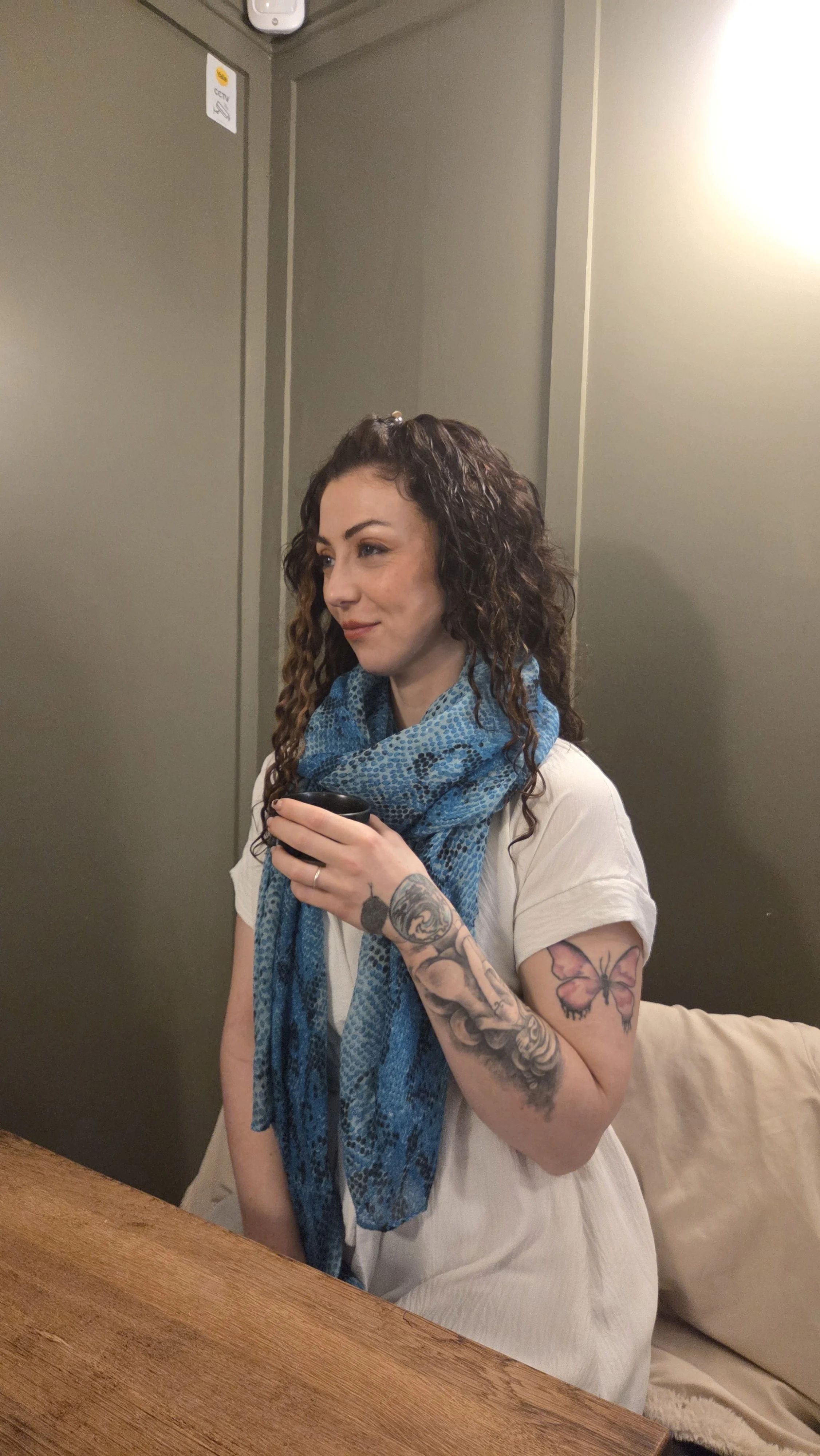 A woman with curly hair, tattoos on her arms, and a blue scarf is standing indoors near a table, holding a phone and smiling slightly.